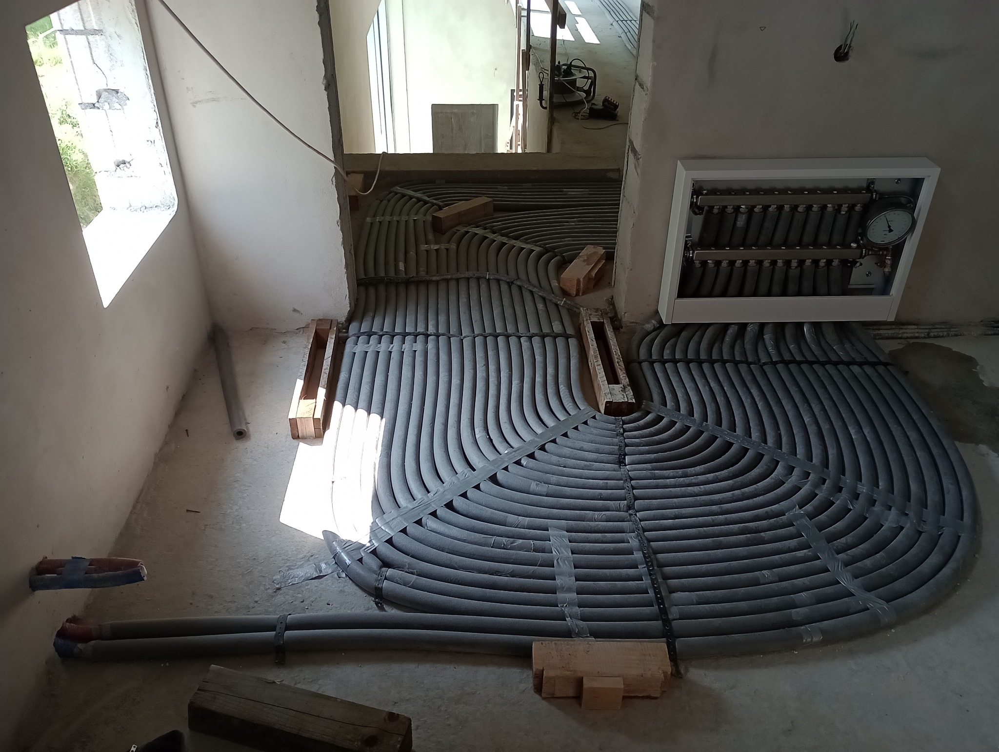 Montaż ogrzewania podłogowego: szare rury ułożone spiralnie na podłodze w surowym stanie, przytwierdzone taśmą, z widocznym rozdzielaczem w białej obudowie na ścianie.