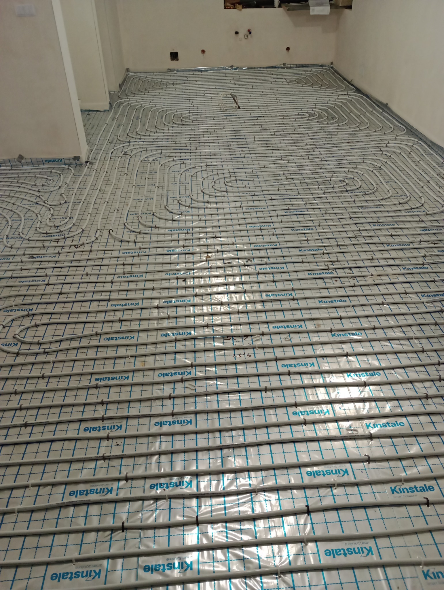 Rozłożona instalacja ogrzewania podłogowego na folii z nadrukiem, widoczne rury w pętlach przed zalaniem wylewką.