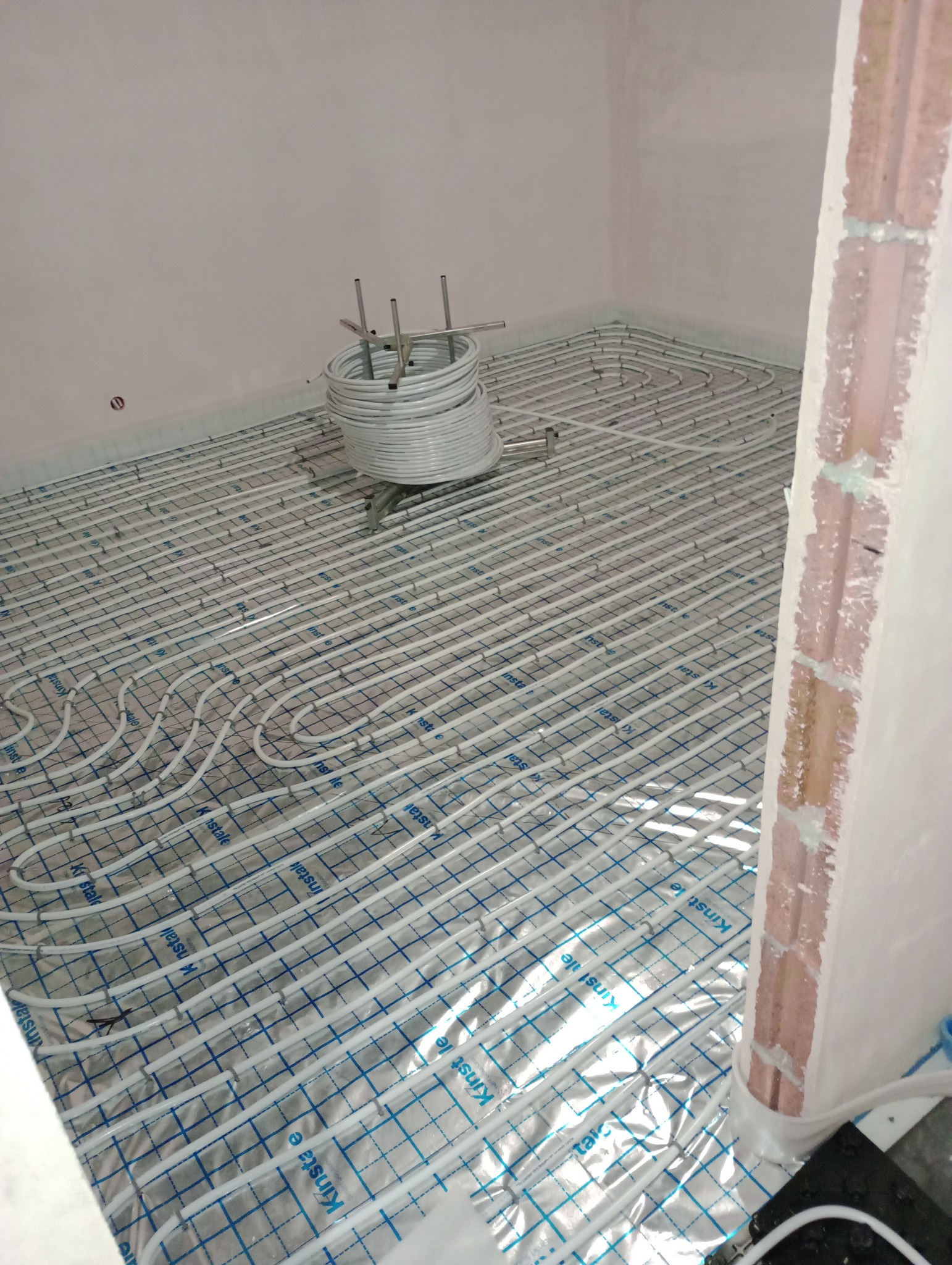 Rozłożone rury ogrzewania podłogowego na folii z nadrukiem, z boku zwinięty zapas rur na metalowym stojaku, surowy stan wykończenia ścian.