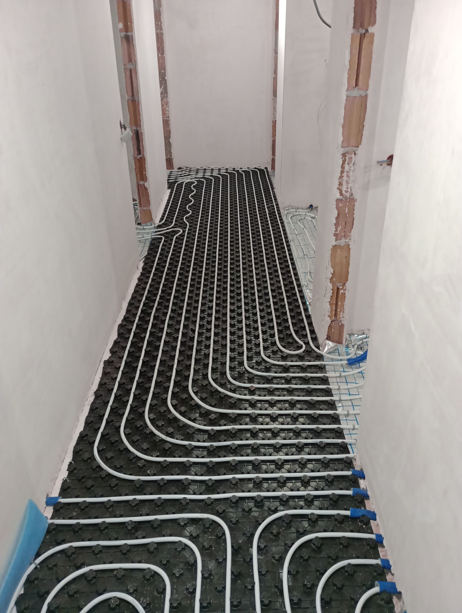 Rozłożone białe rury ogrzewania podłogowego na czarnej macie izolacyjnej w trakcie montażu, widoczne ściany w surowym stanie i elementy instalacji.
