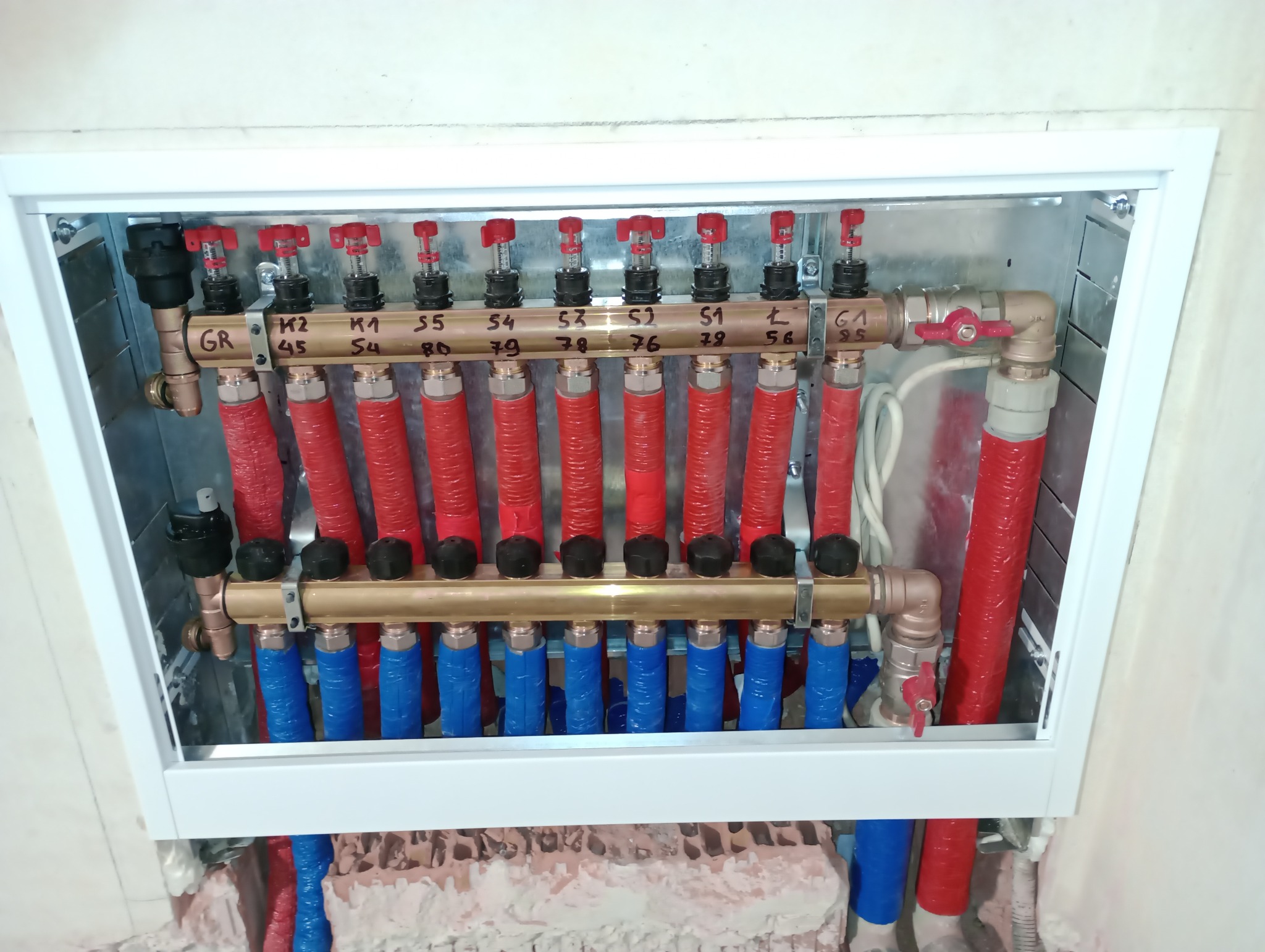 Rozdzielacz ogrzewania podłogowego w metalowej szafce z numerowanymi zaworami i rurami w izolacji czerwonej i niebieskiej, widok z góry.