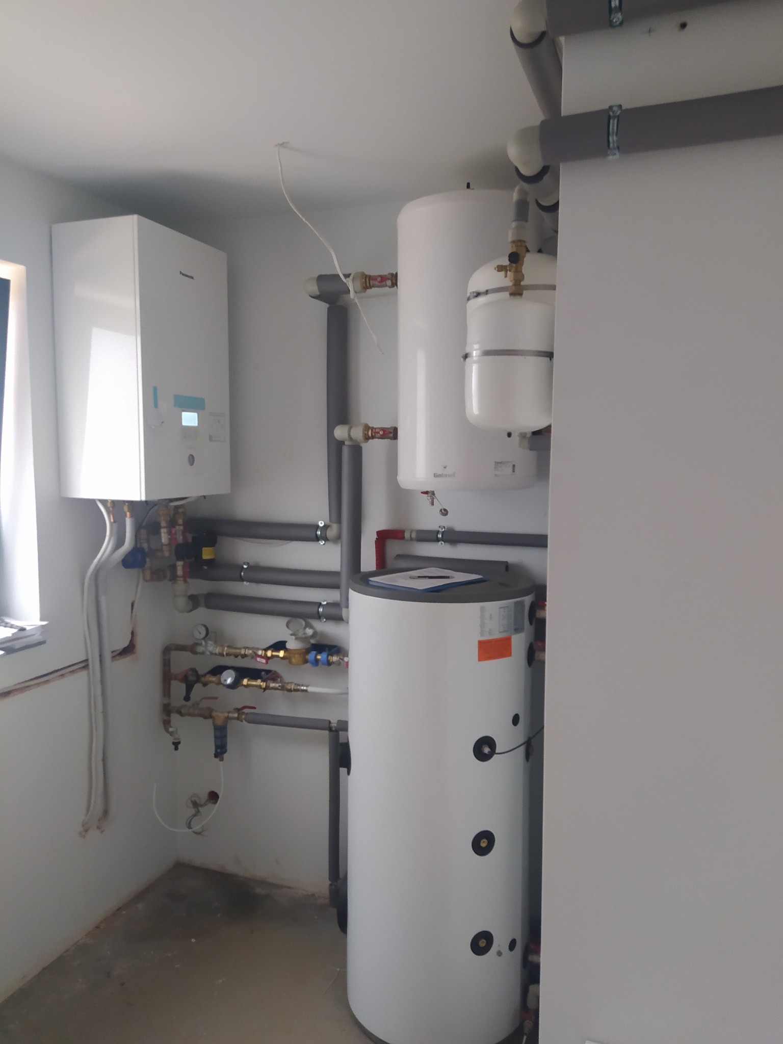 Wnętrze kotłowni z zamontowanym kondensacyjnym kotłem gazowym Panasonic, zasobnikiem ciepłej wody użytkowej Galmet i instalacją hydrauliczną z rur stalowych.
