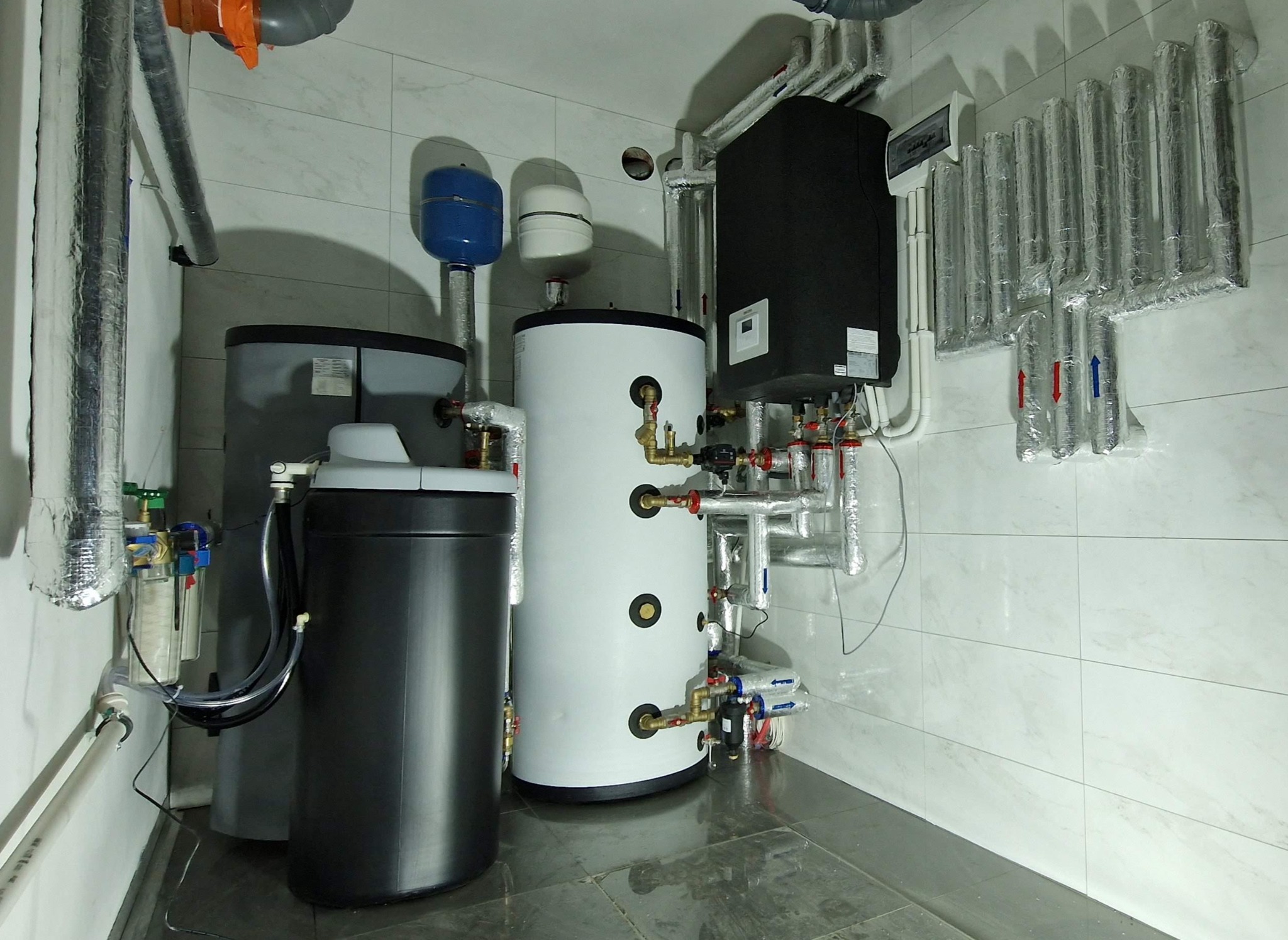 Kotłownia z zasobnikami wody, instalacją grzewczą i filtrami, widok na białe kafelki ścienne i szarą podłogę.