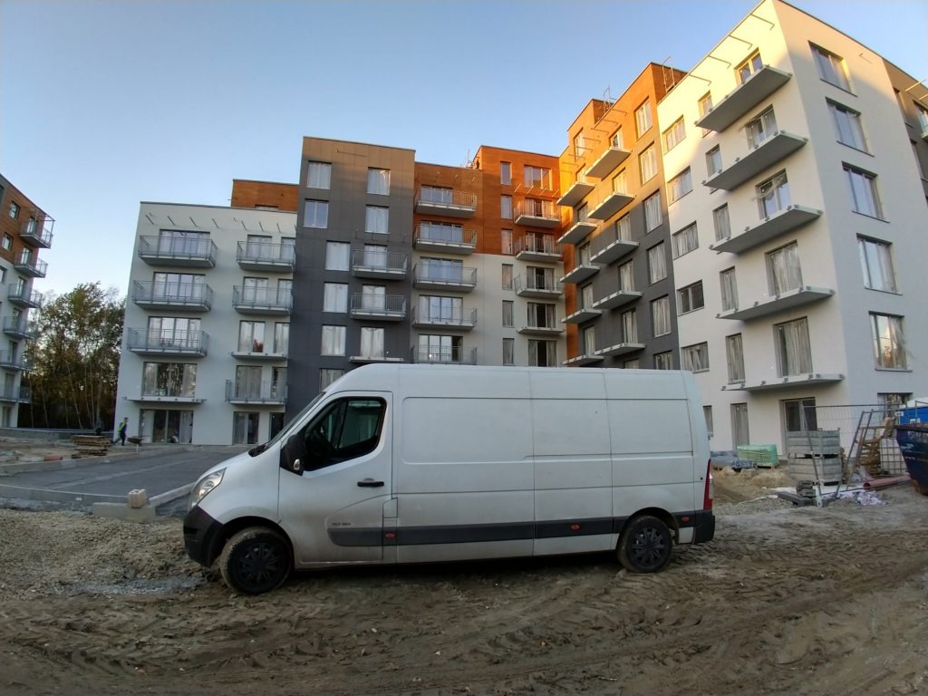 Biały furgonet zaparkowany na placu budowy przed nowoczesnym budynkiem mieszkalnym z balkonami, widoczne ślady błota na oponach i karoserii.