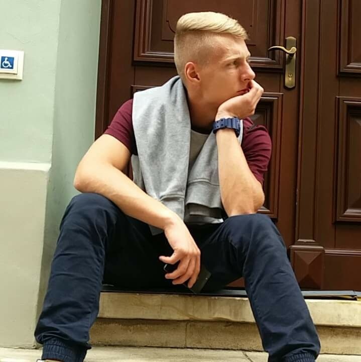 Młody mężczyzna siedzi na schodach przed brązowymi drzwiami, zamyślony, w tle widoczny znak dla niepełnosprawnych na zielonej ścianie.