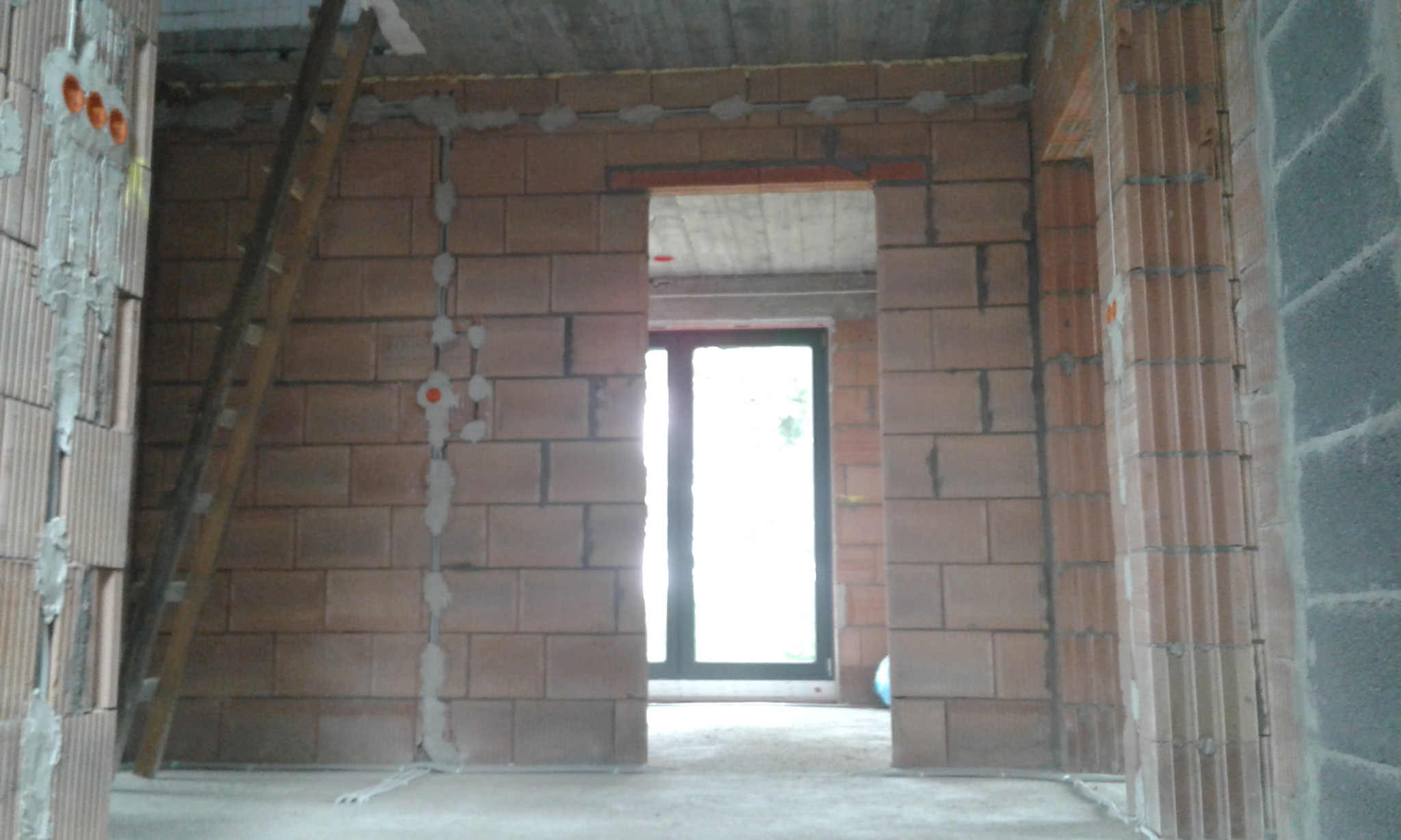 Wnętrze surowego budynku w trakcie prac, widoczne bruzdy pod kable elektryczne w ścianach z cegły, drabina oparta o ścianę, otwór drzwiowy z widokiem na dalszą część budynku.