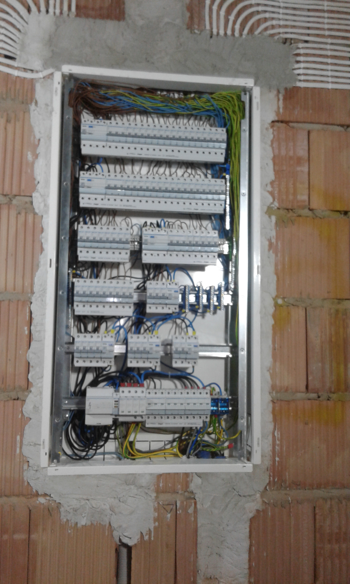 Rozdzielnica elektryczna z licznymi bezpiecznikami zamontowana w surowej ścianie z cegły, widoczne kable w różnych kolorach i rurki instalacyjne w ścianie.