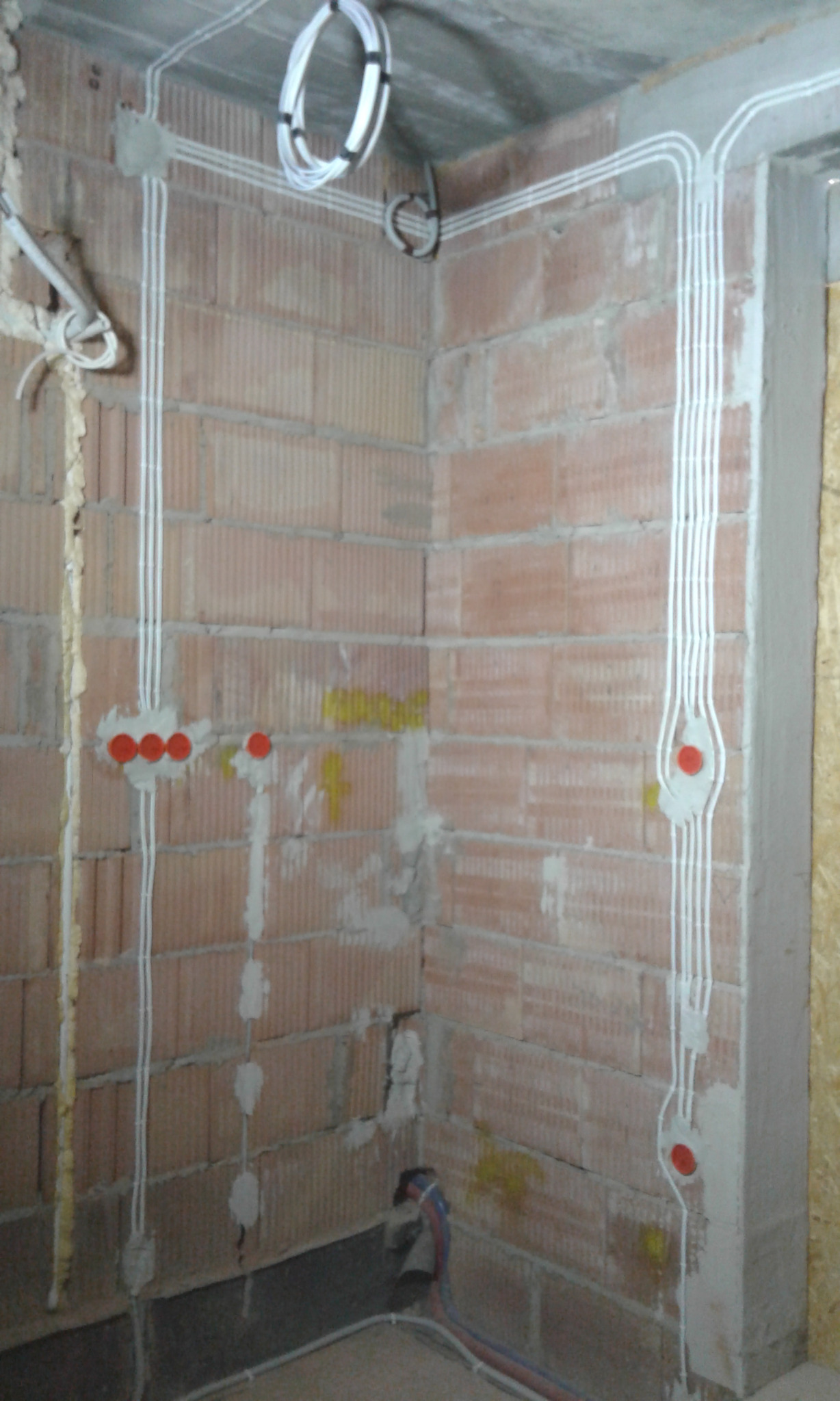 Surowe ściany z cegły dziurawki z rozprowadzonymi przewodami elektrycznymi w białych peszlach, widoczne puszki instalacyjne z czerwonymi zaślepkami, oraz zwinięte kable pod sufitem.