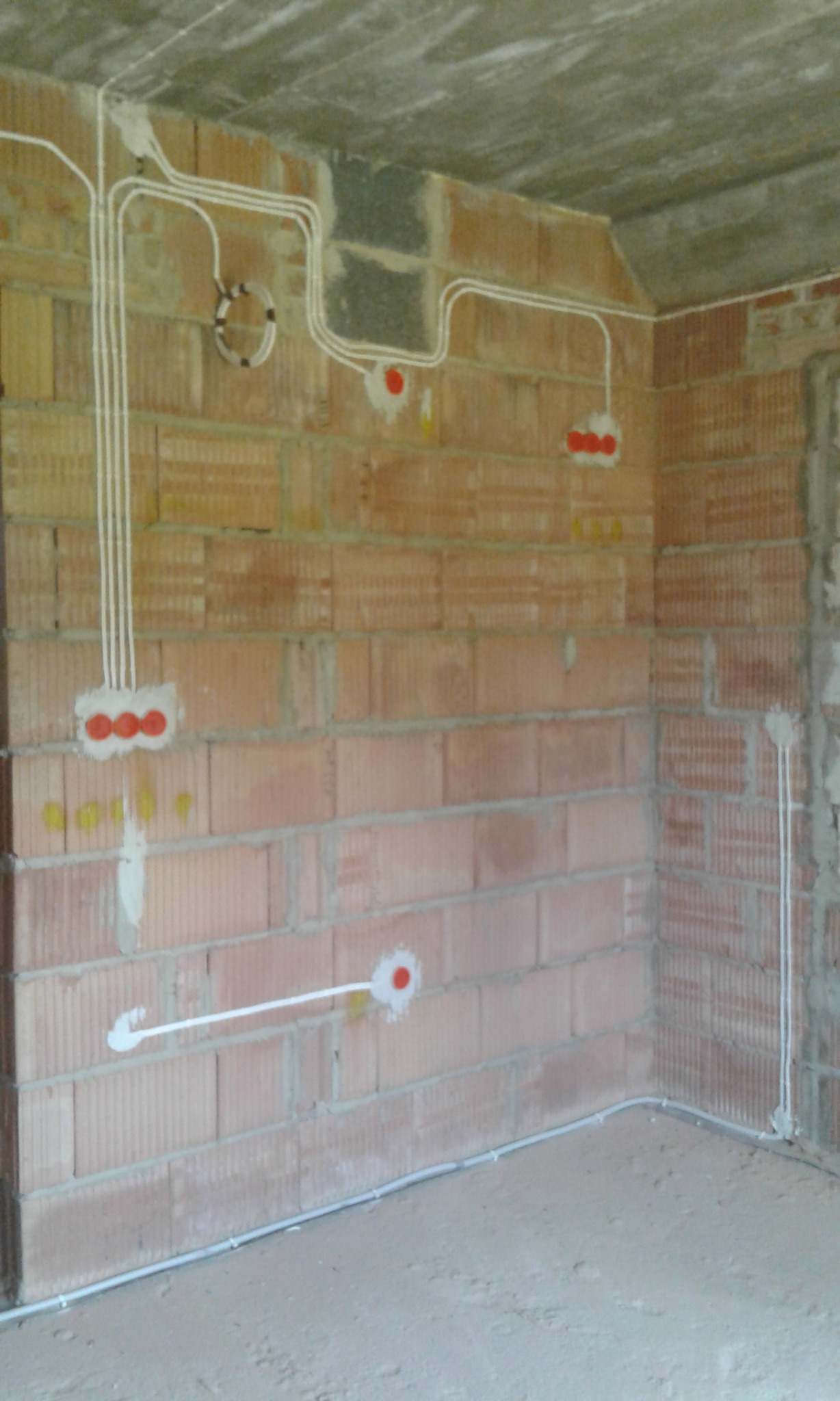 Surowy mur z cegły dziurawki z widocznymi, nowo poprowadzonymi przewodami elektrycznymi w białych rurkach, prowadzącymi do puszek instalacyjnych z czerwonymi zaślepkami. Część przewodów zwinięta...