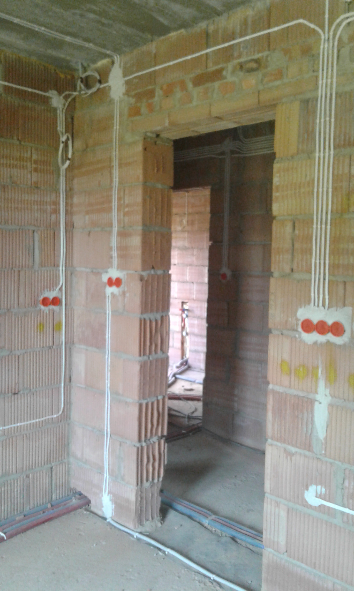 Wnętrze w stanie surowym z widocznymi przewodami elektrycznymi poprowadzonymi po ścianach z cegły, puszki instalacyjne z czerwonymi zaślepkami, przewody ułożone wzdłuż podłogi.