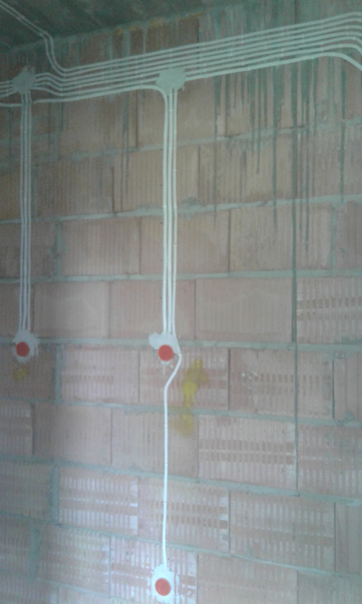 Białe kable elektryczne prowadzone natynkowo po ceglanej ścianie w surowym stanie, widoczne puszki instalacyjne z czerwonymi zaślepkami.