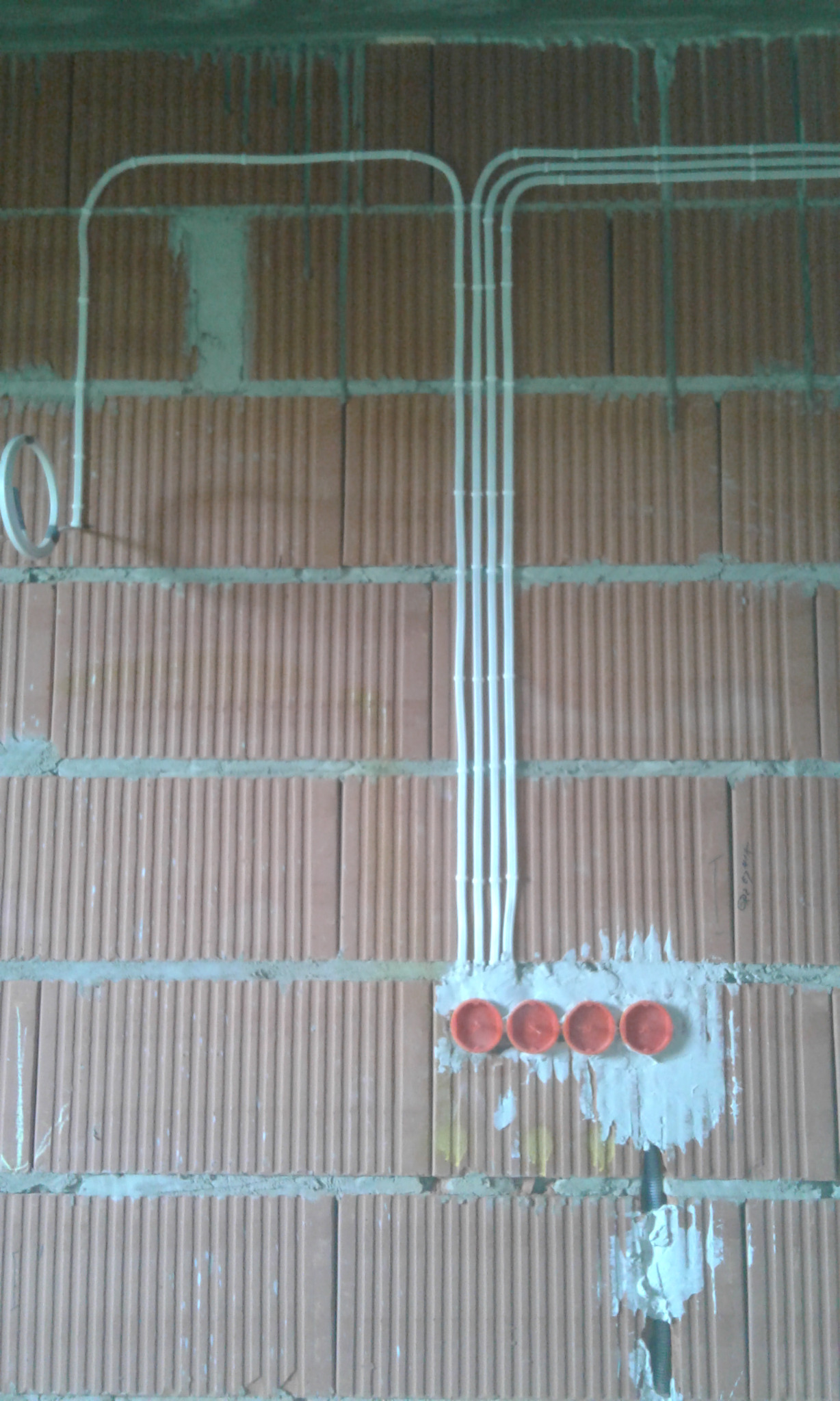 Białe rurki instalacji elektrycznej ułożone na ścianie z cegły, zabezpieczone pomarańczowymi osłonkami, widoczne ślady zaprawy.