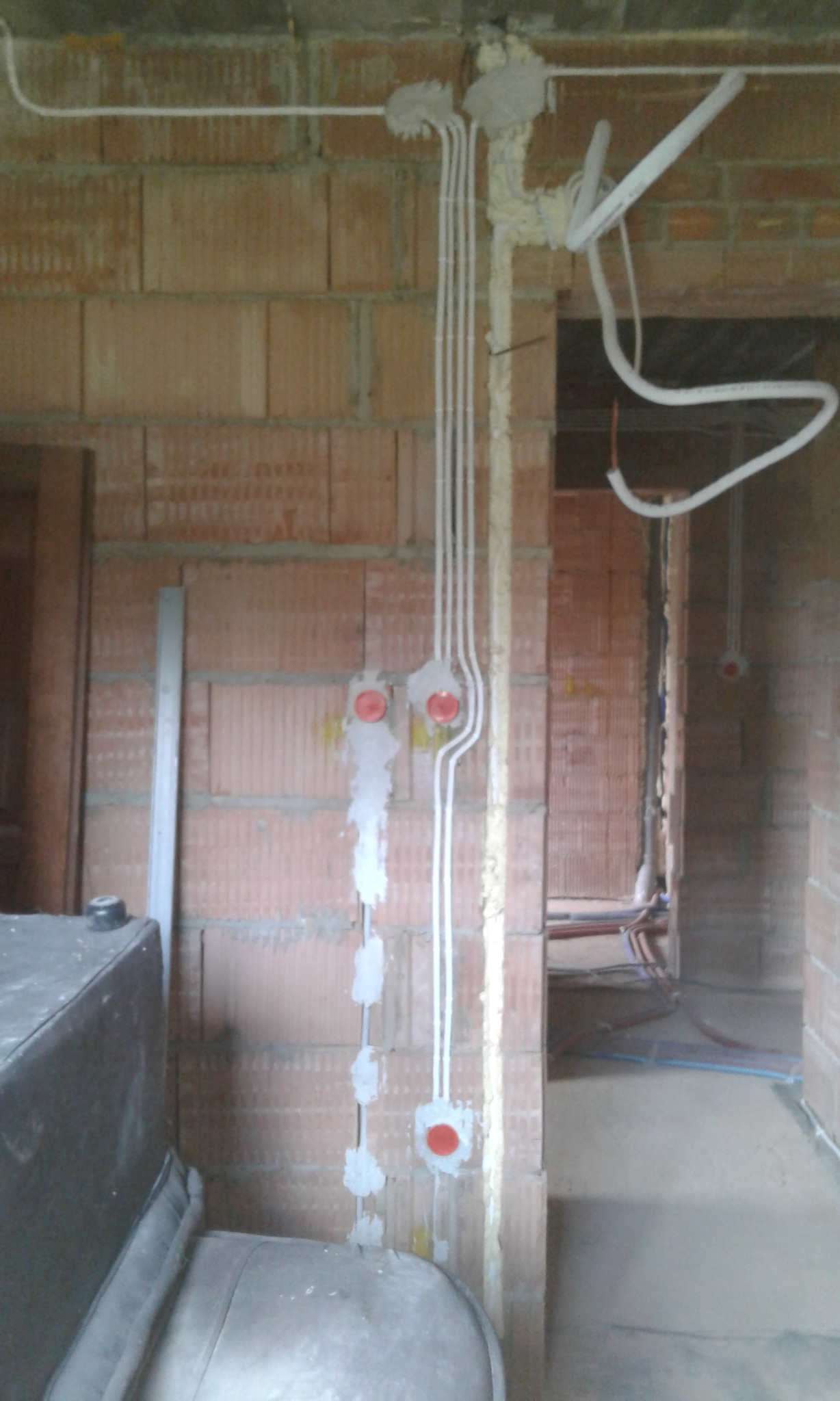 Surowe ściany z pustaków z widocznymi przewodami elektrycznymi w białych rurkach, zamocowanymi pianką montażową, z pomarańczowymi puszkami instalacyjnymi, w tle widoczne instalacje wodne.