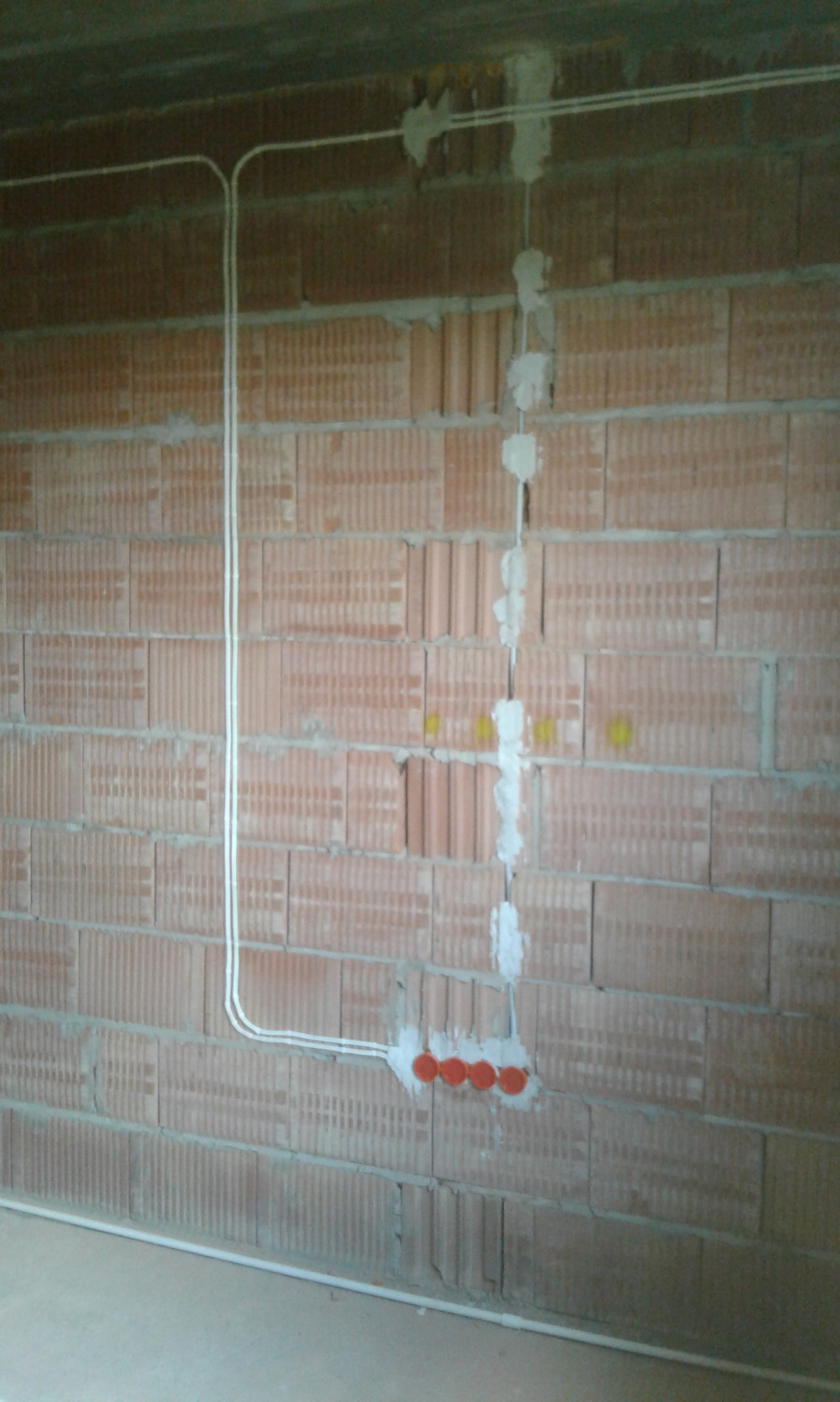 Instalacja elektryczna w trakcie montażu na ścianie z cegły dziurawki, widoczne białe przewody w peszlu, puszki elektryczne z pomarańczowymi zaślepkami i gipsowe mocowania.