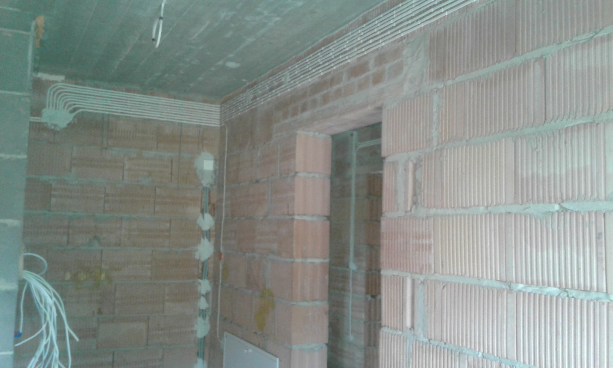 Wnętrze w trakcie budowy z widocznymi przewodami elektrycznymi ułożonymi pod sufitem i wzdłuż ściany, zamocowanymi do surowych, czerwonych cegieł.