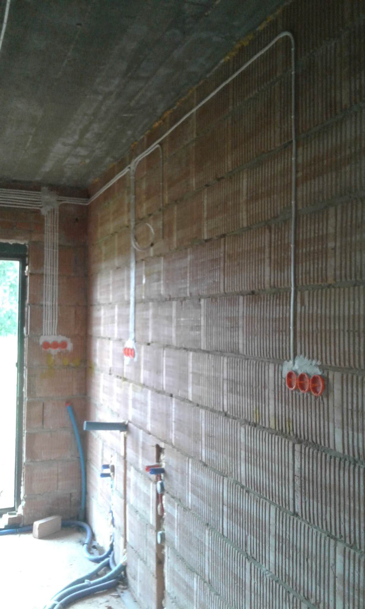 Surowe wnętrze w trakcie prac wykończeniowych: widoczne kable elektryczne prowadzone po ścianie z cegły ceramicznej, puszki instalacyjne w kolorze pomarańczowym, fragment okna po lewej stronie...