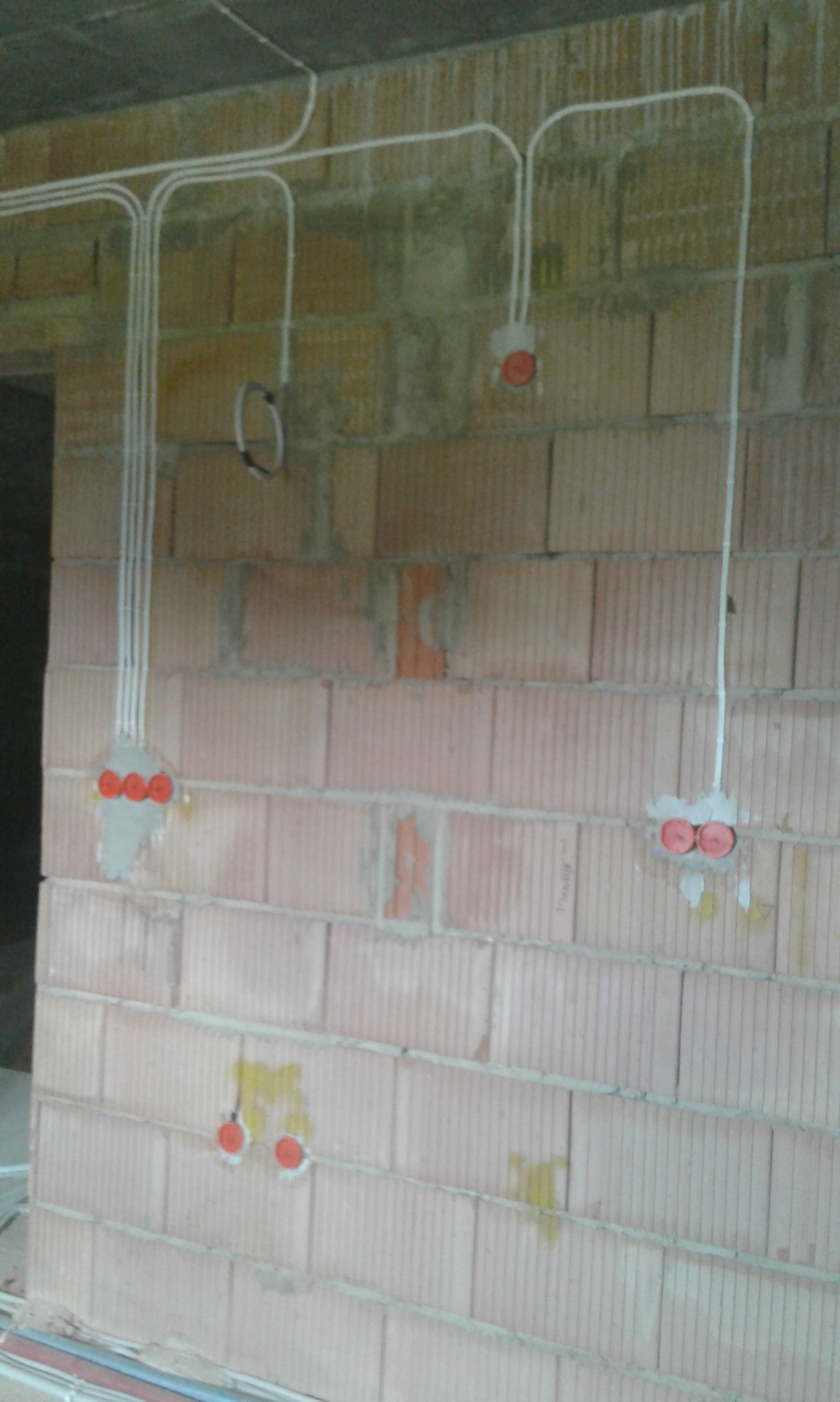 Świeżo poprowadzone białe przewody elektryczne w peszlach na surowej ścianie z cegły dziurawki, widoczne puszki instalacyjne z czerwonymi zaślepkami, przygotowane do osadzenia gniazdek.