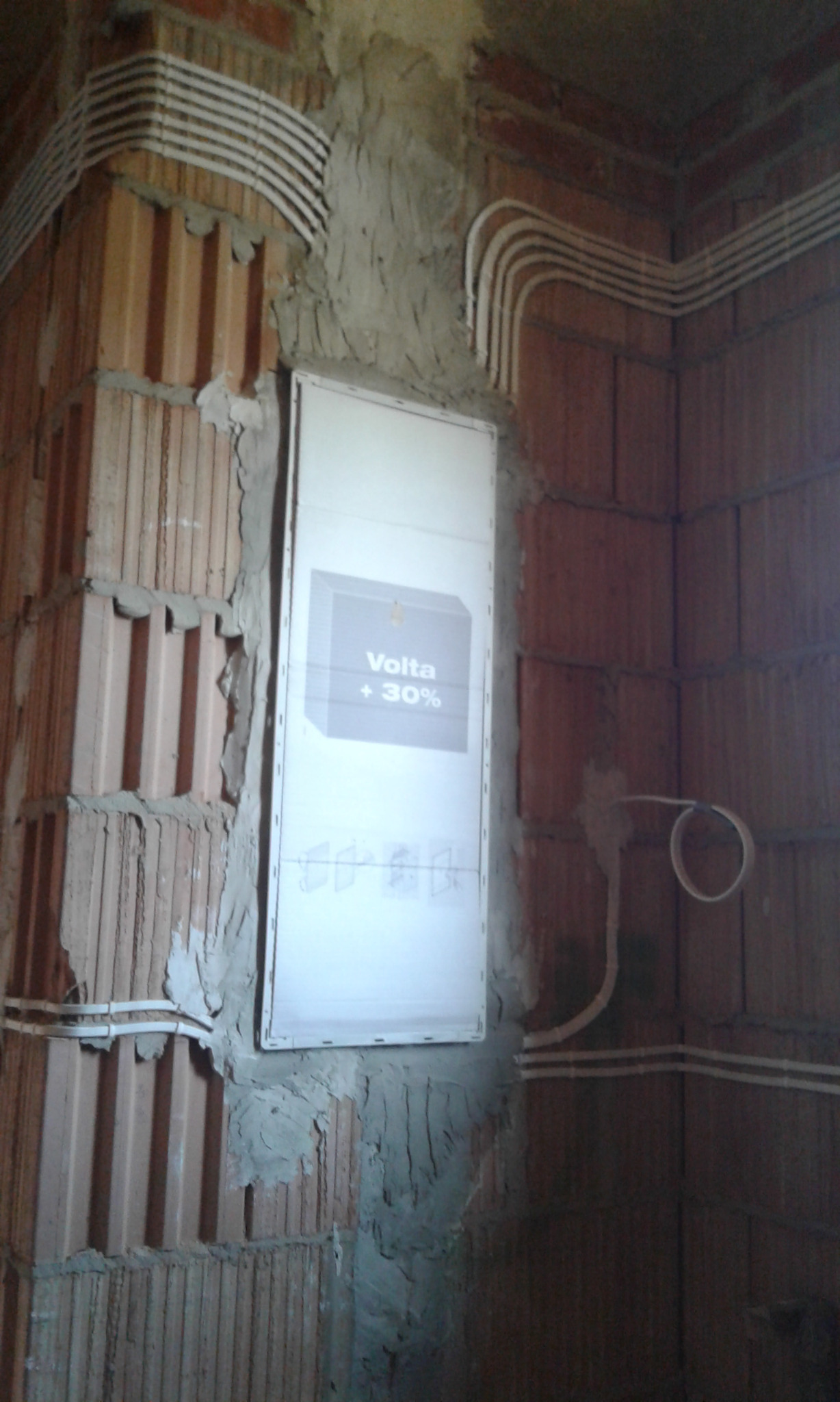 Rozdzielnica elektryczna Volta +30% zamontowana na surowej ścianie z cegły dziurawki, z widocznymi przewodami elektrycznymi w białych peszlach. Ściana w trakcie prac wykończeniowych, z widocznymi...