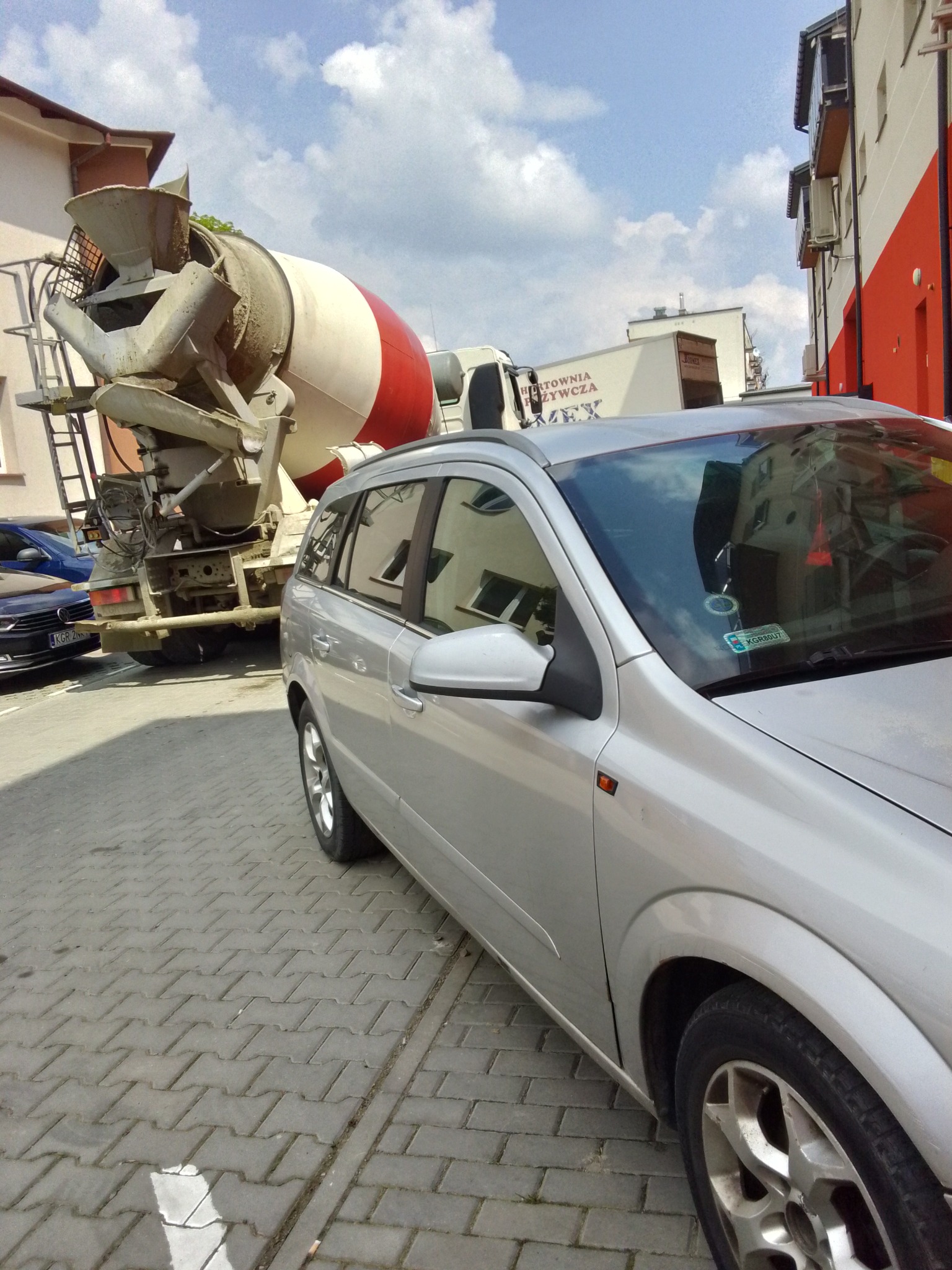 Ciężarówka z betoniarką zaparkowana na brukowanej ulicy, obok srebrnego samochodu osobowego, w tle budynki mieszkalne i błękitne niebo z chmurami.