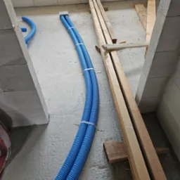 Dwa niebieskie karbowane przewody ułożone równolegle na surowym betonowym podłożu w przestrzeni między ścianami z bloczków, zabezpieczone metalowymi uchwytami, obok leżą drewniane belki...