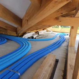 Niebieskie rury wentylacyjne ułożone na poddaszu w trakcie montażu, widoczne drewniane elementy konstrukcji dachu.