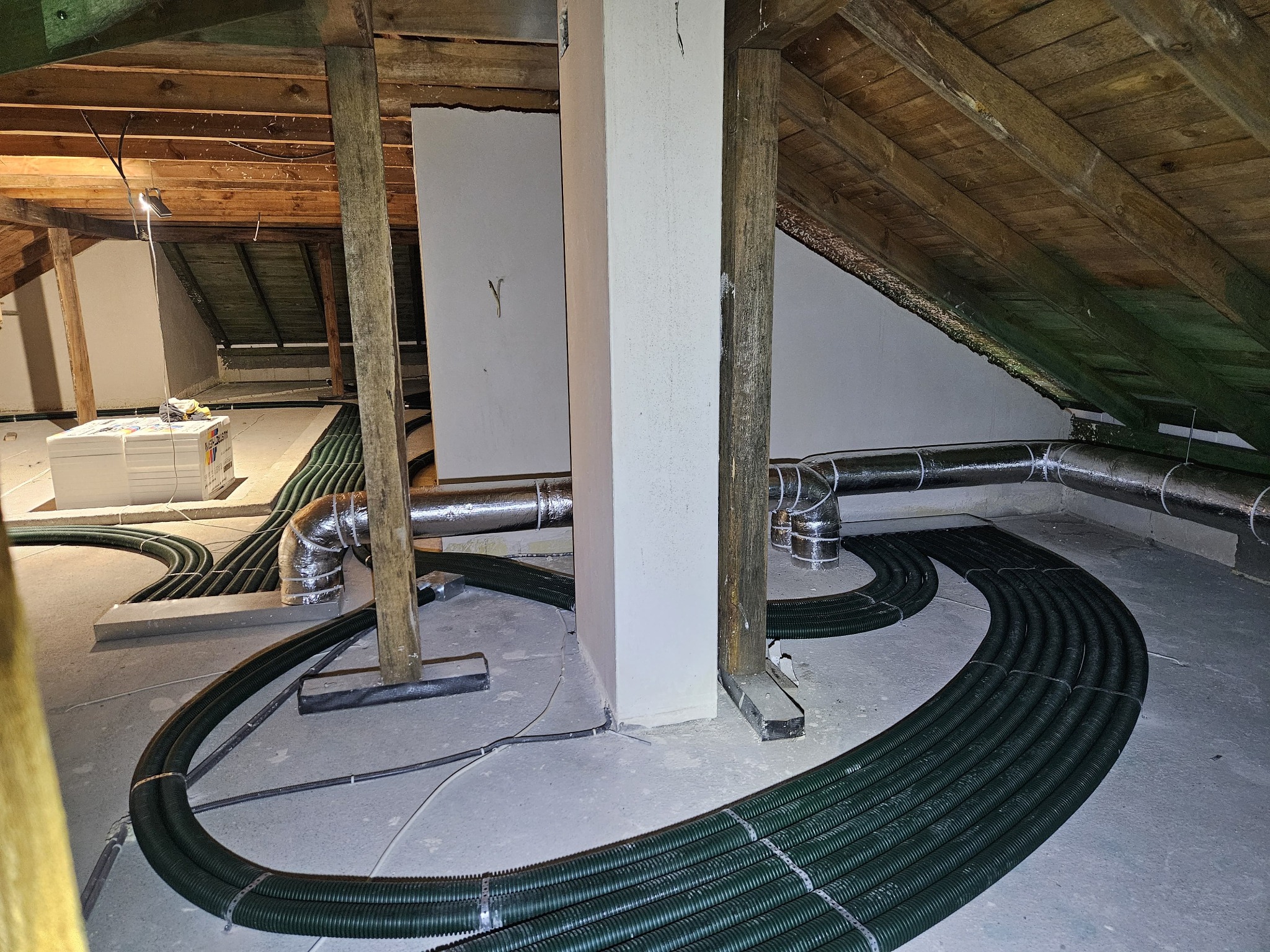 Montaż instalacji wentylacyjnej z zielonymi rurami w surowym wnętrzu poddasza z drewnianą konstrukcją dachu i białą ścianą.