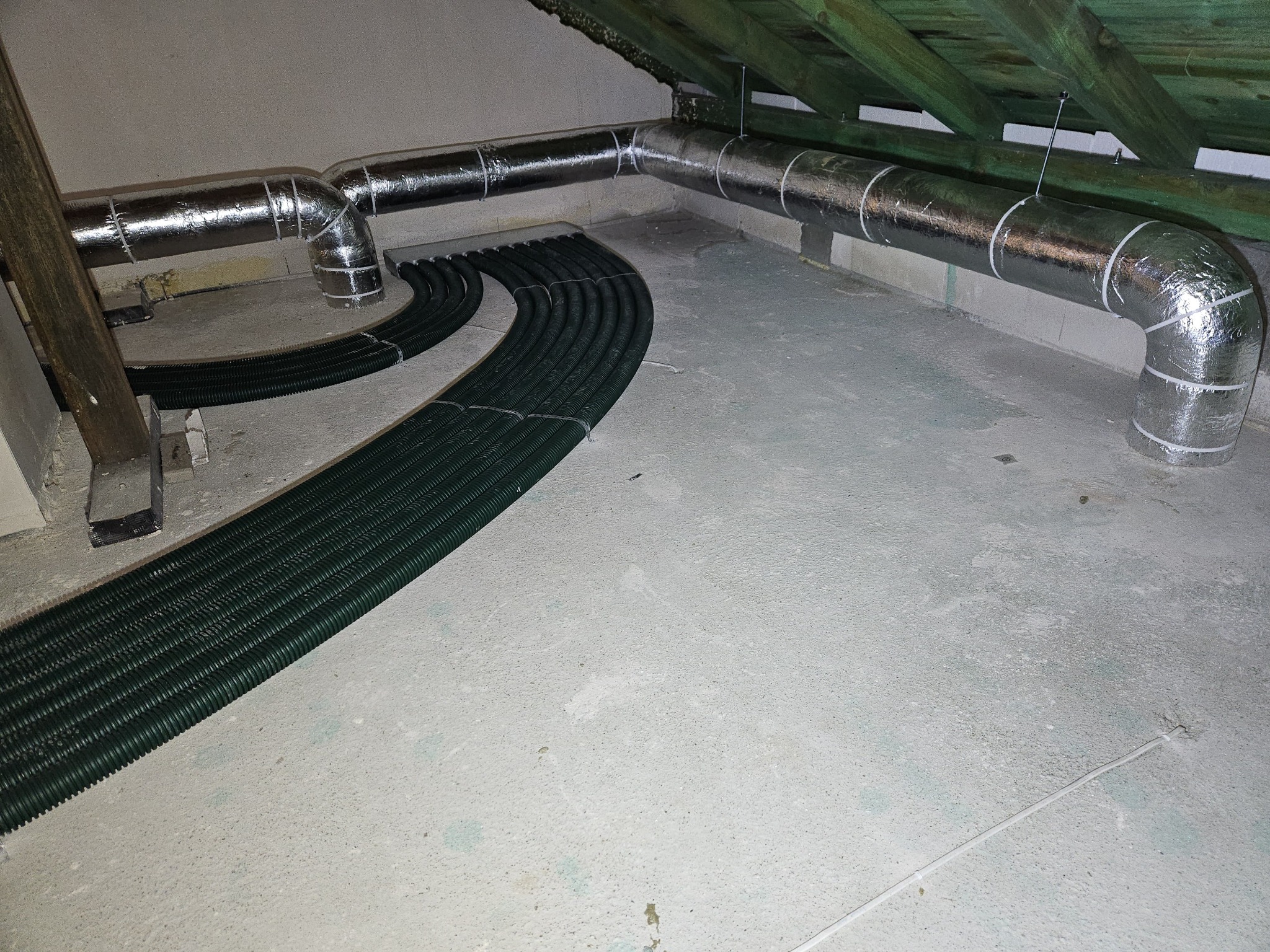Szare, izolowane rury wentylacyjne ułożone pod sufitem na poddaszu, z zielonymi, karbowanymi przewodami ułożonymi równolegle na betonowej podłodze.