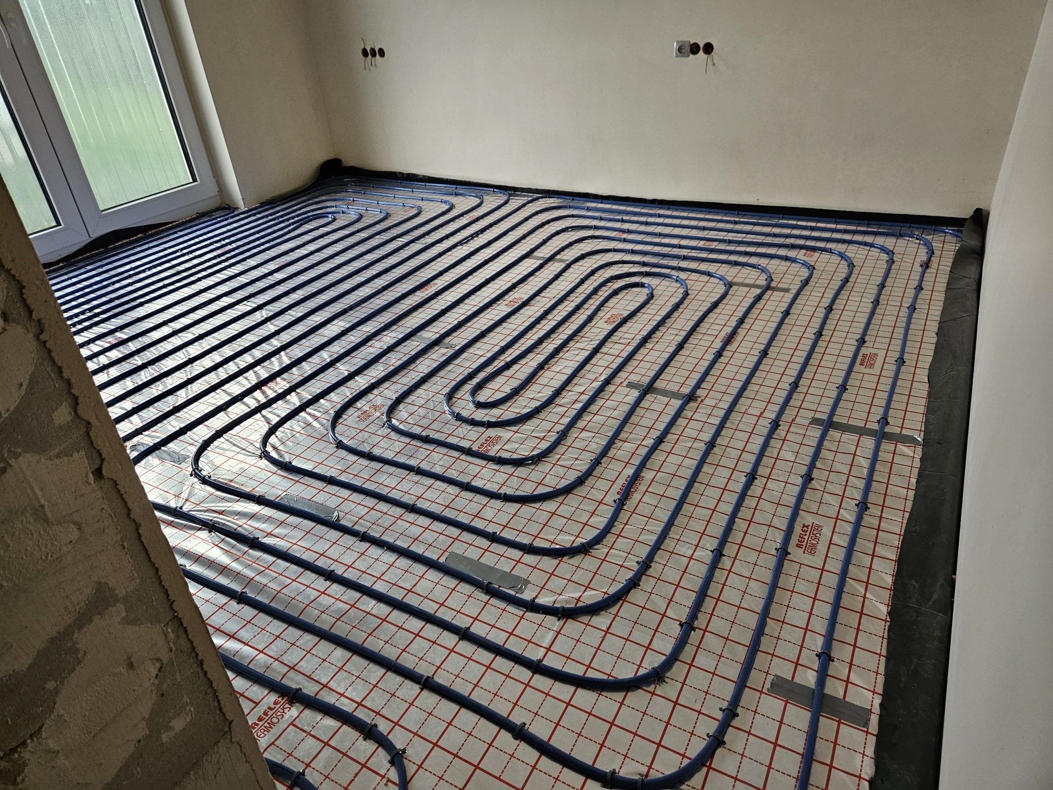 Niebieskie rury ogrzewania podłogowego ułożone spiralnie na folii z czerwoną siatką, widoczne mocowania i taśma, surowy stan wykończenia wnętrza z oknem i wystającymi przewodami elektrycznymi.