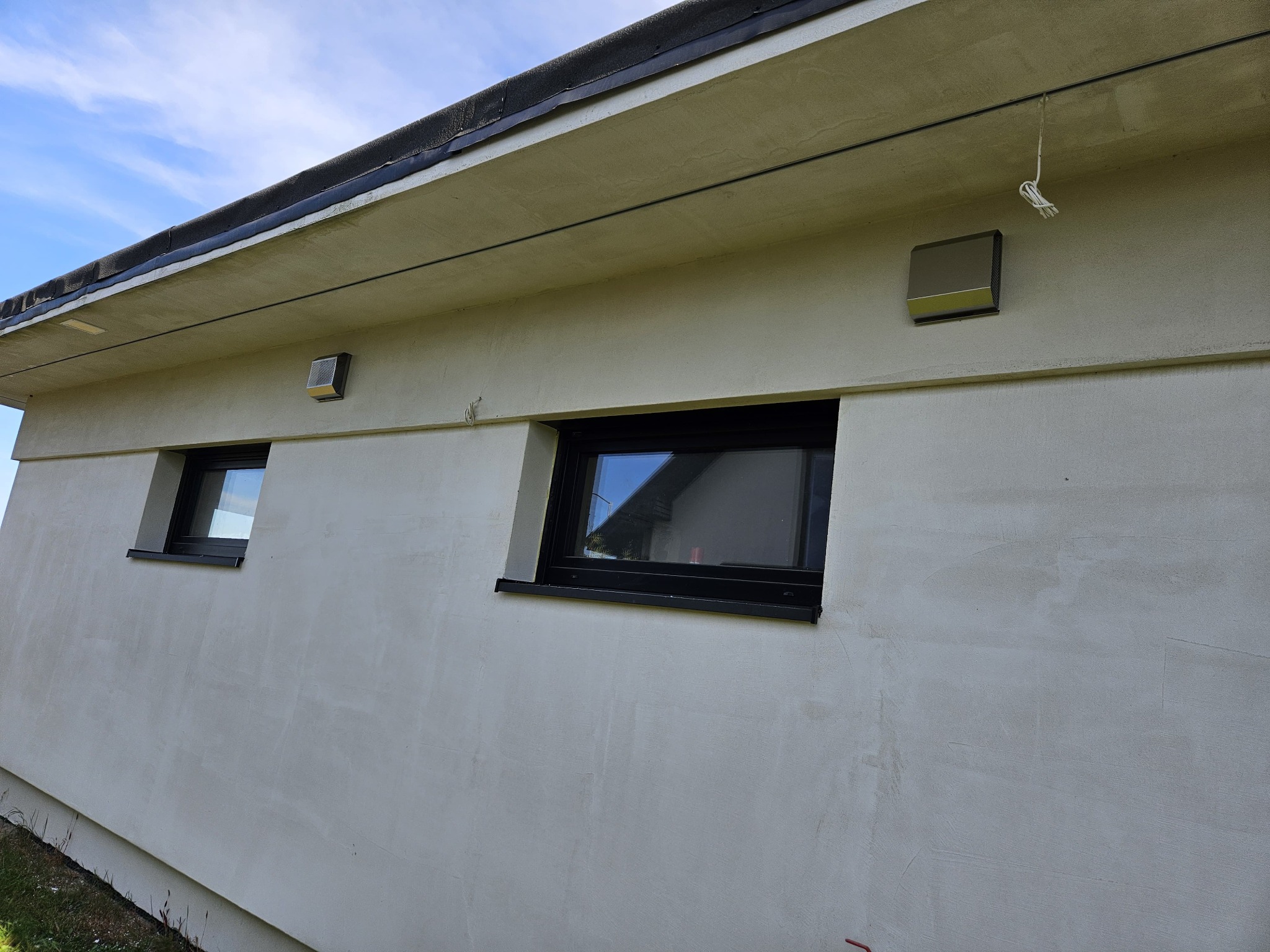 Zewnętrzna ściana budynku z dwoma czarnymi oknami i szarymi kratkami wentylacyjnymi pod okapem, widoczne niedoskonałości tynku.