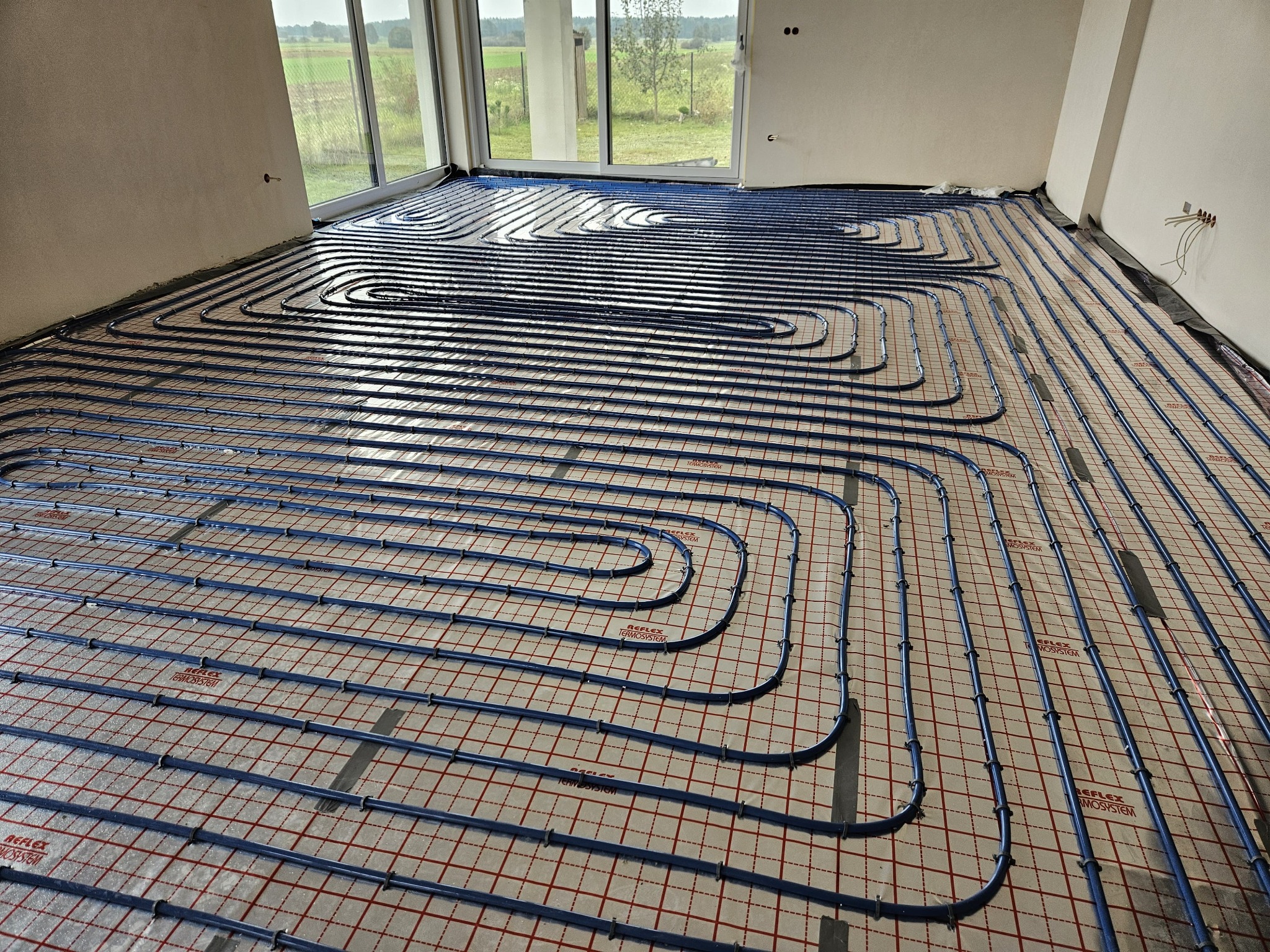 Rozłożone niebieskie rury ogrzewania podłogowego na folii z czerwoną siatką w surowym wnętrzu z dużym oknem wychodzącym na zielony krajobraz.