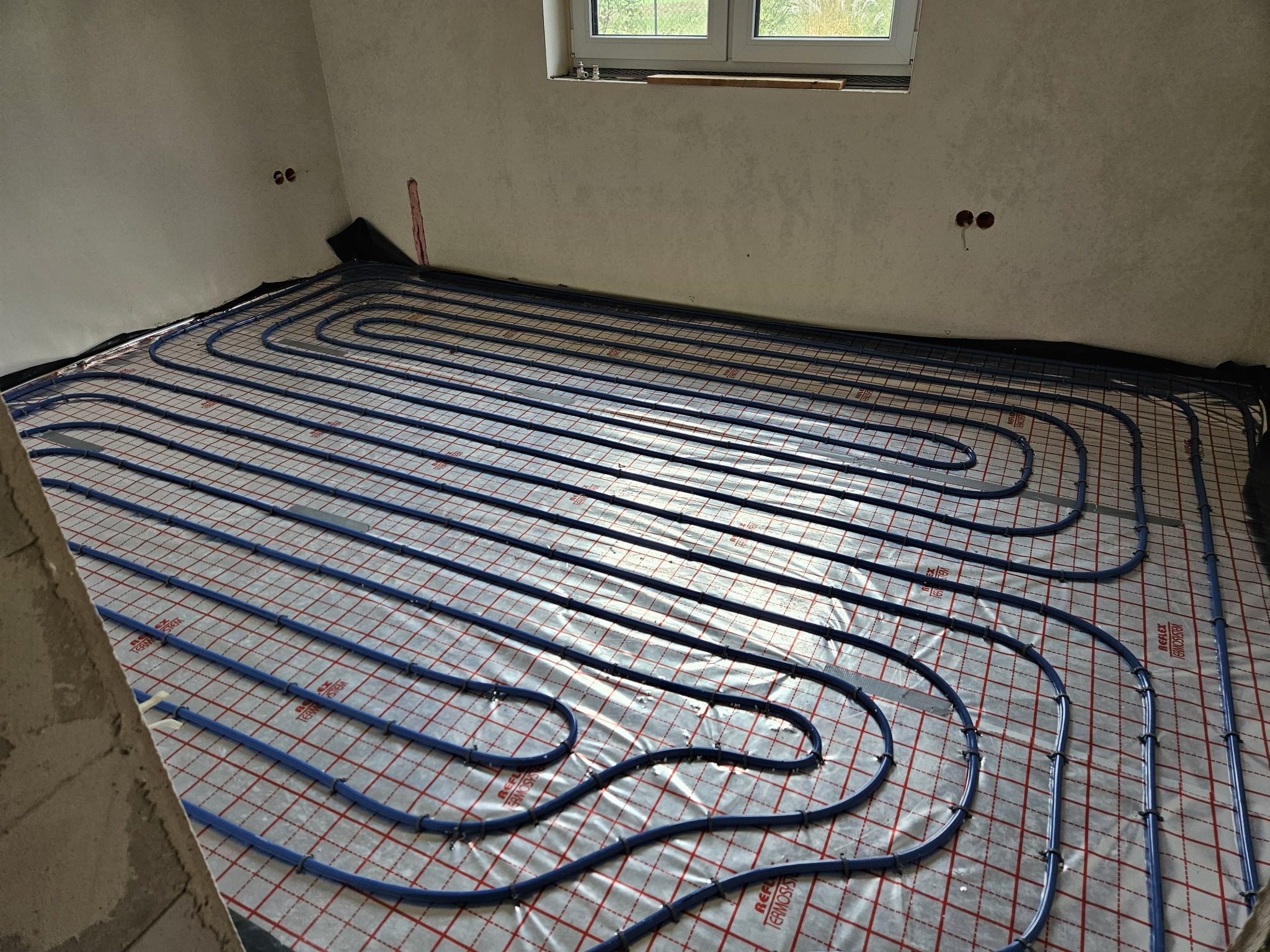 Niebieskie rury ogrzewania podłogowego ułożone na folii z czerwoną siatką w surowym pomieszczeniu z oknem.
