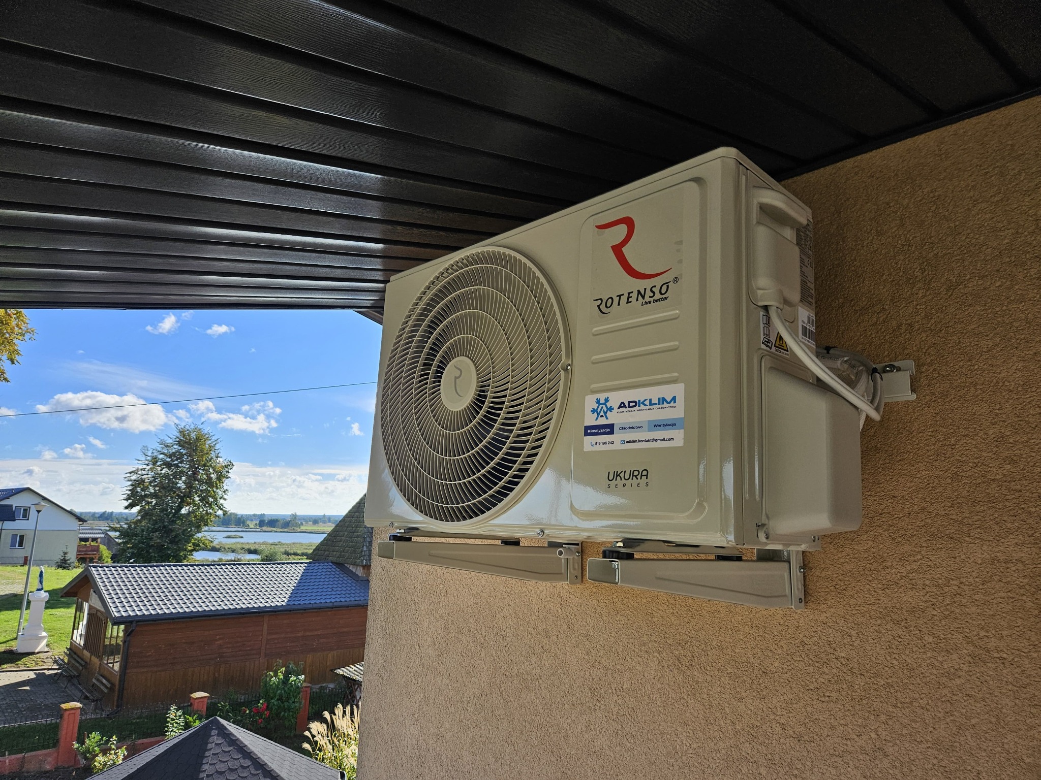 Zewnętrzna jednostka klimatyzacji Rotenso Ukura Series zamontowana na elewacji budynku z widokiem na krajobraz wiejski.