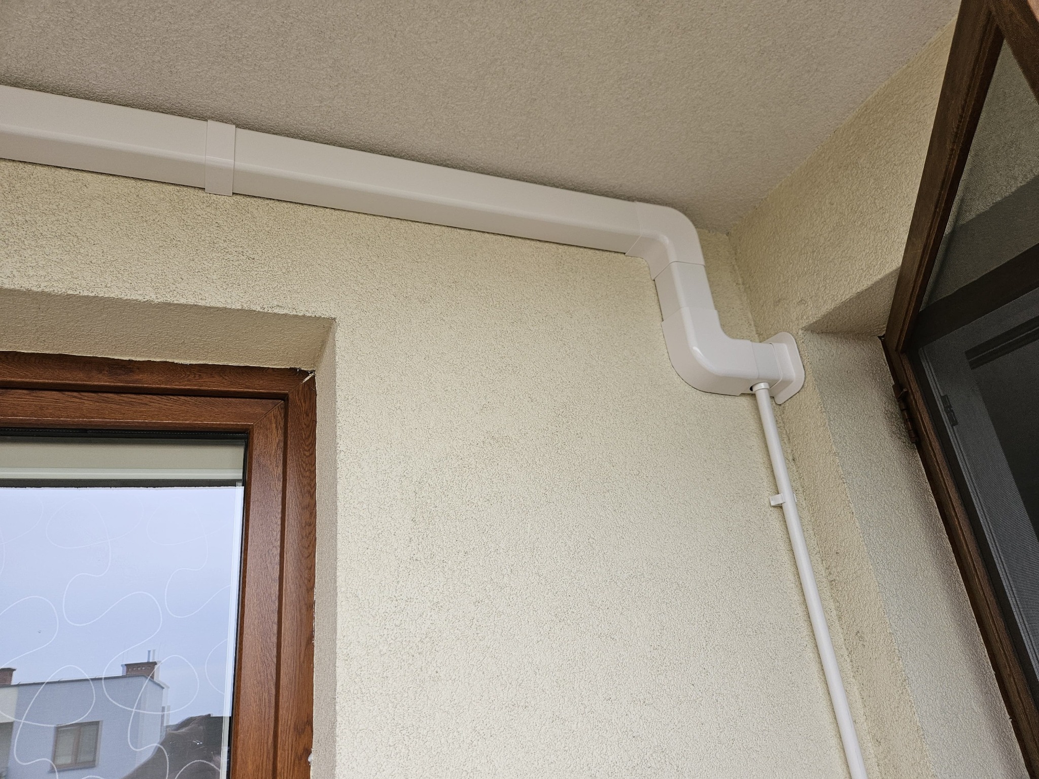 Białe, plastikowe kanały wentylacyjne przymocowane do ściany z fakturą, z widocznym oknem z brązową ramą i siatką.