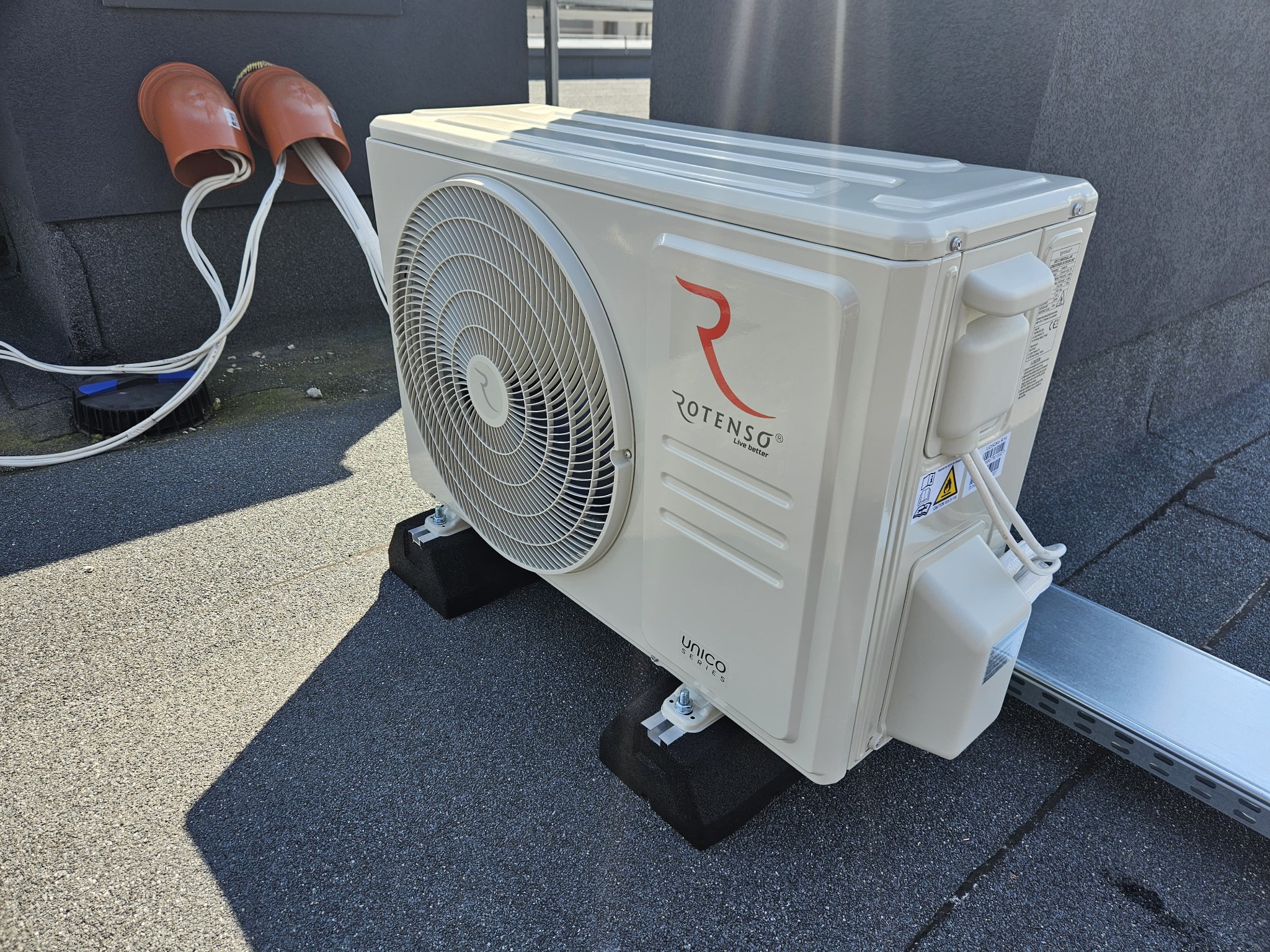 Zewnętrzna jednostka klimatyzacji Rotenso Unico Series zamontowana na dachu, widoczne przewody wchodzące do budynku przez pomarańczowe rury ochronne.