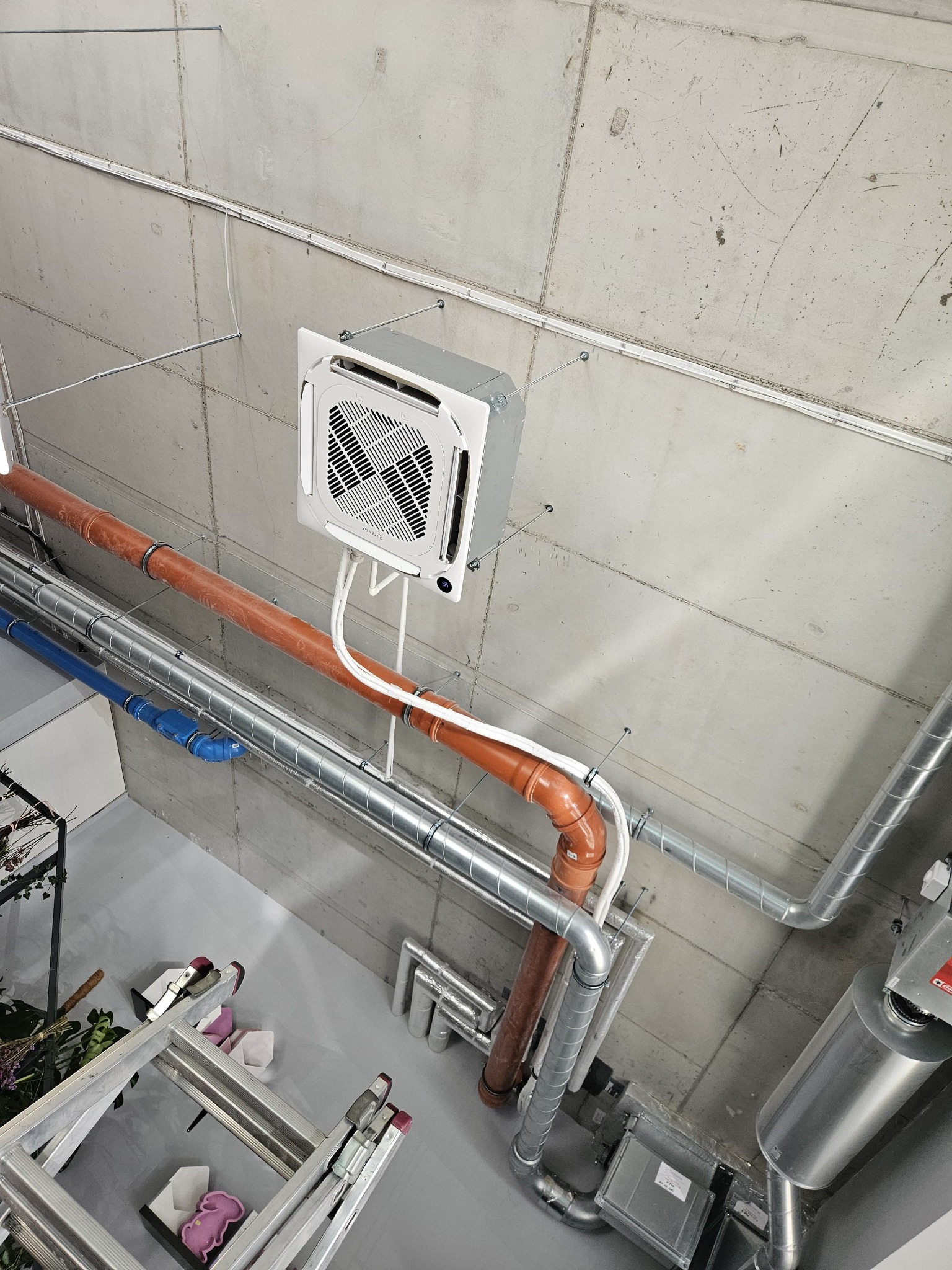 Sufit z zamontowaną kasetą klimatyzacyjną, widoczne rury wentylacyjne (spiro) i instalacja hydrauliczna, drabina aluminiowa z narzędziami na podłodze.