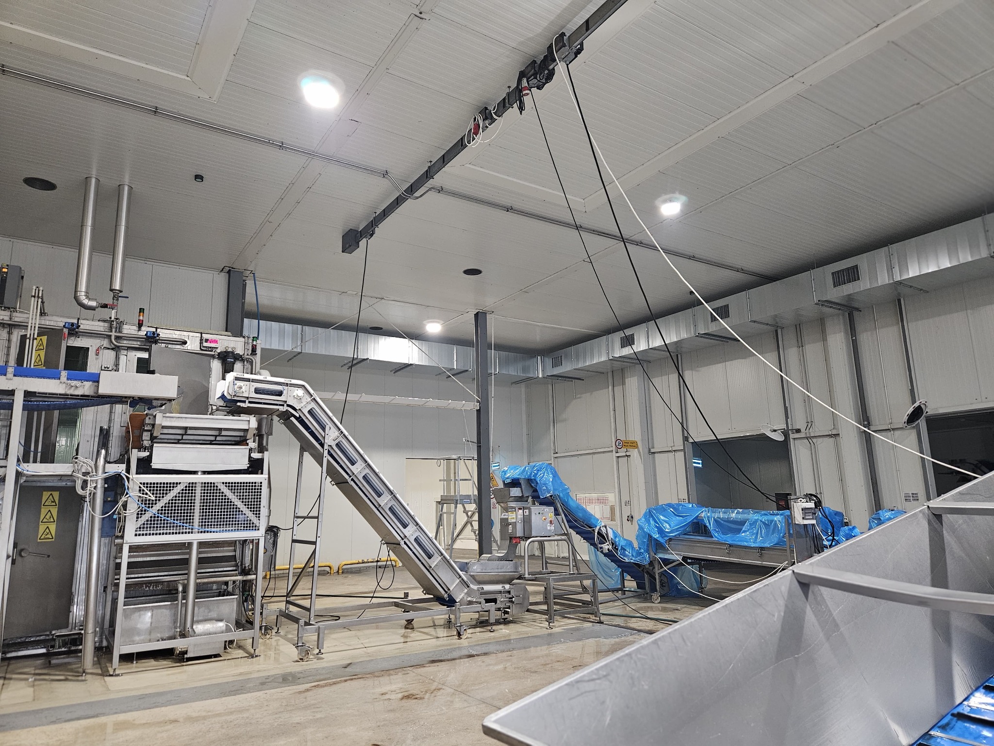 Wnętrze hali produkcyjnej z systemem transportu taśmowego, widoczne elementy wentylacji pod sufitem i instalacje hydrauliczne przy maszynach.