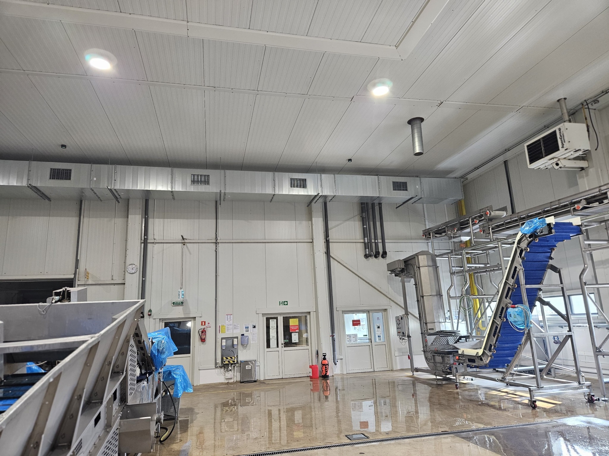 Wnętrze hali produkcyjnej z systemem wentylacji i klimatyzacji, widoczne kanały wentylacyjne pod sufitem, urządzenia chłodnicze oraz linia produkcyjna z taśmociągiem i elementami ze stali...