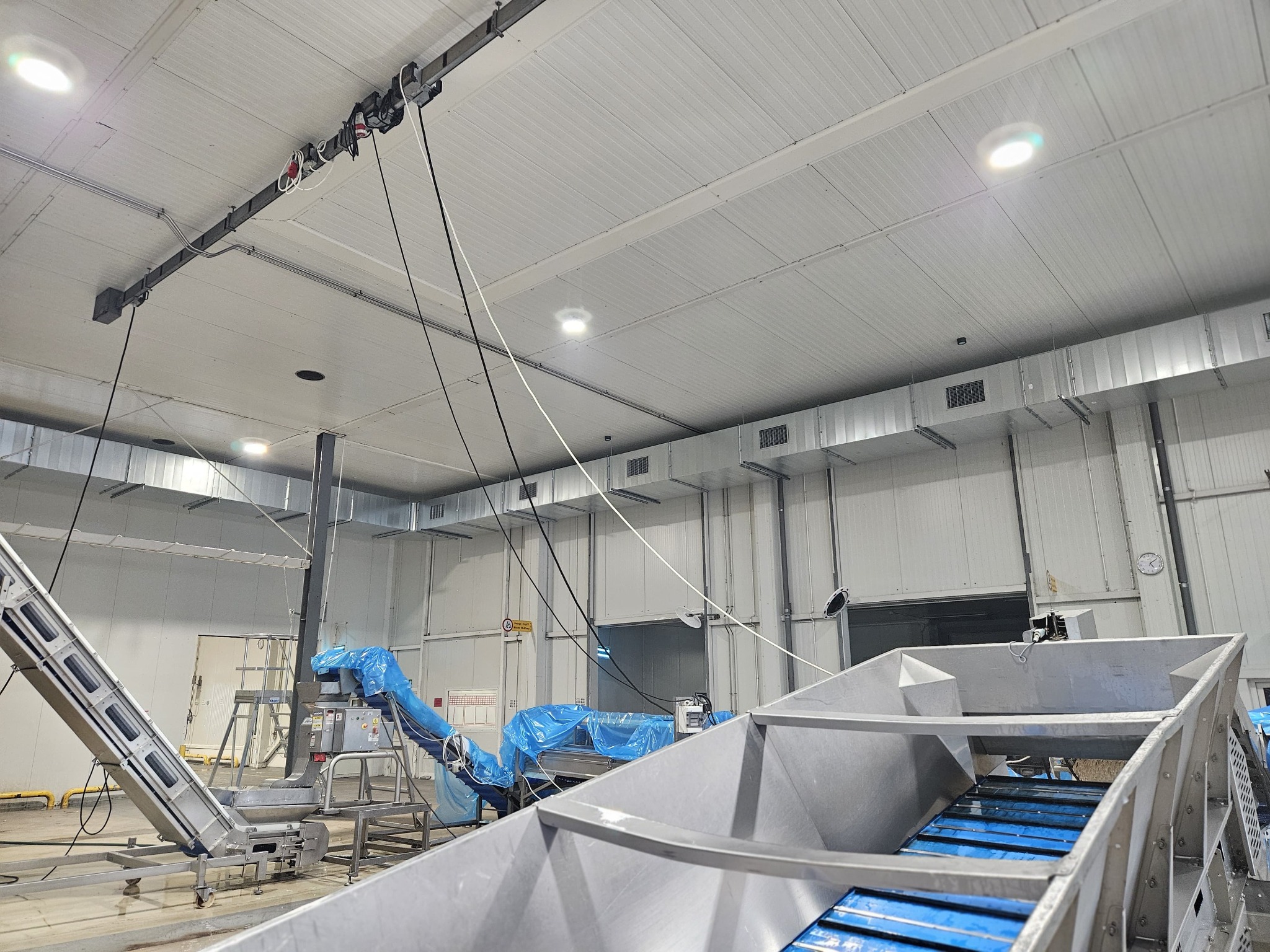 Wnętrze hali produkcyjnej z widocznymi elementami systemu wentylacji pod sufitem oraz fragmentami linii produkcyjnej z transporterami taśmowymi.