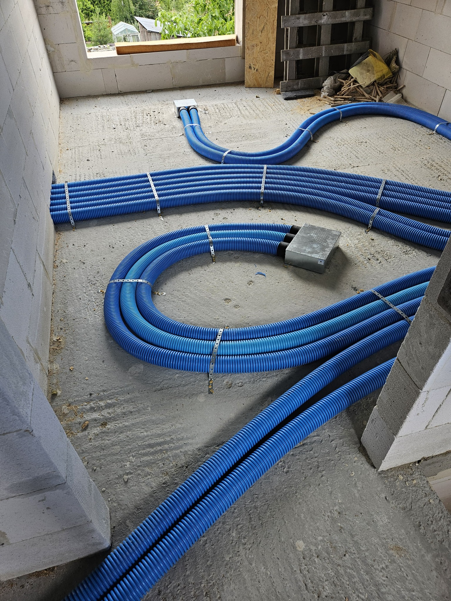 Niebieskie rury karbowane, spięte metalowymi klamrami, ułożone na surowym betonowym podłożu w trakcie instalacji w budynku z białych bloczków.