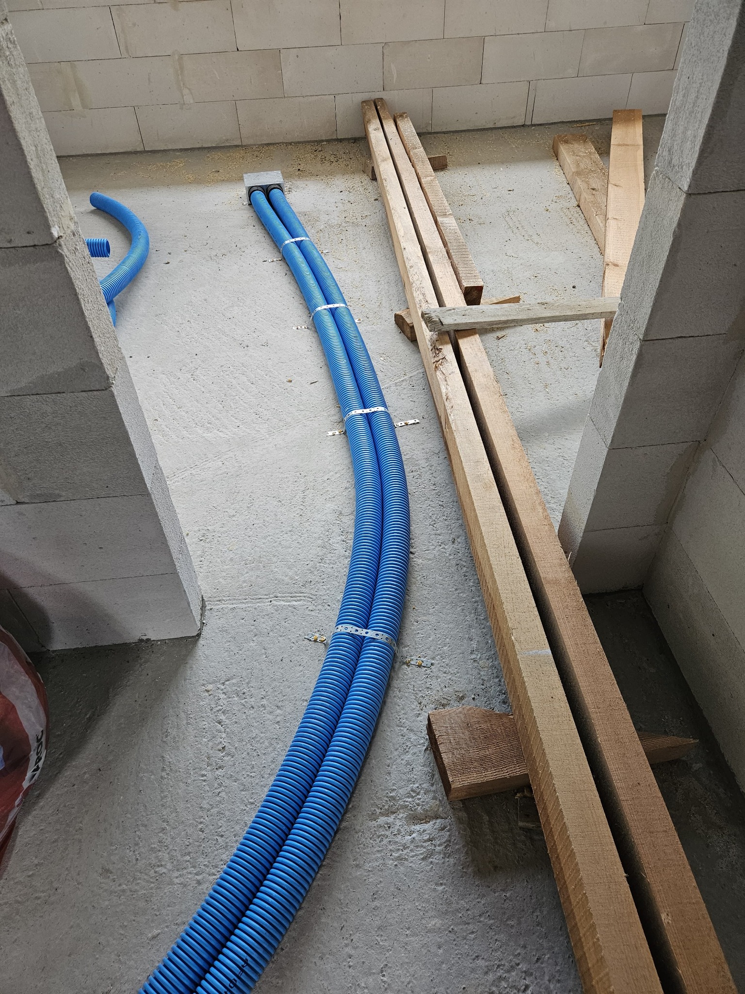 Dwa niebieskie karbowane przewody ułożone równolegle na surowym betonowym podłożu w przestrzeni między ścianami z bloczków, zabezpieczone metalowymi uchwytami, obok leżą drewniane belki...