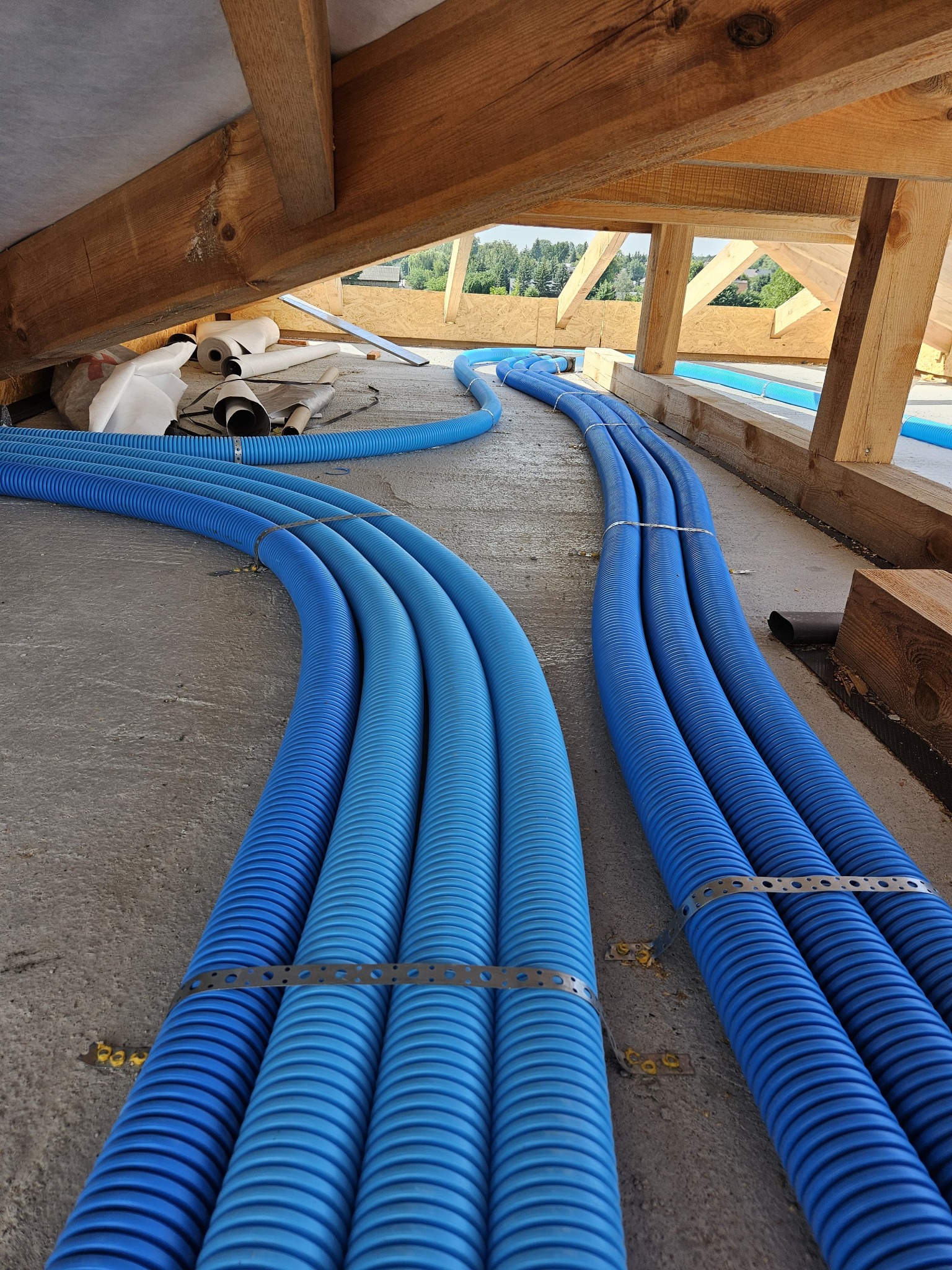 Niebieskie, karbowane rury wentylacyjne ułożone równolegle na surowym podłożu poddasza, zabezpieczone metalowymi taśmami montażowymi, z widoczną drewnianą konstrukcją dachu i fragmentem okna...