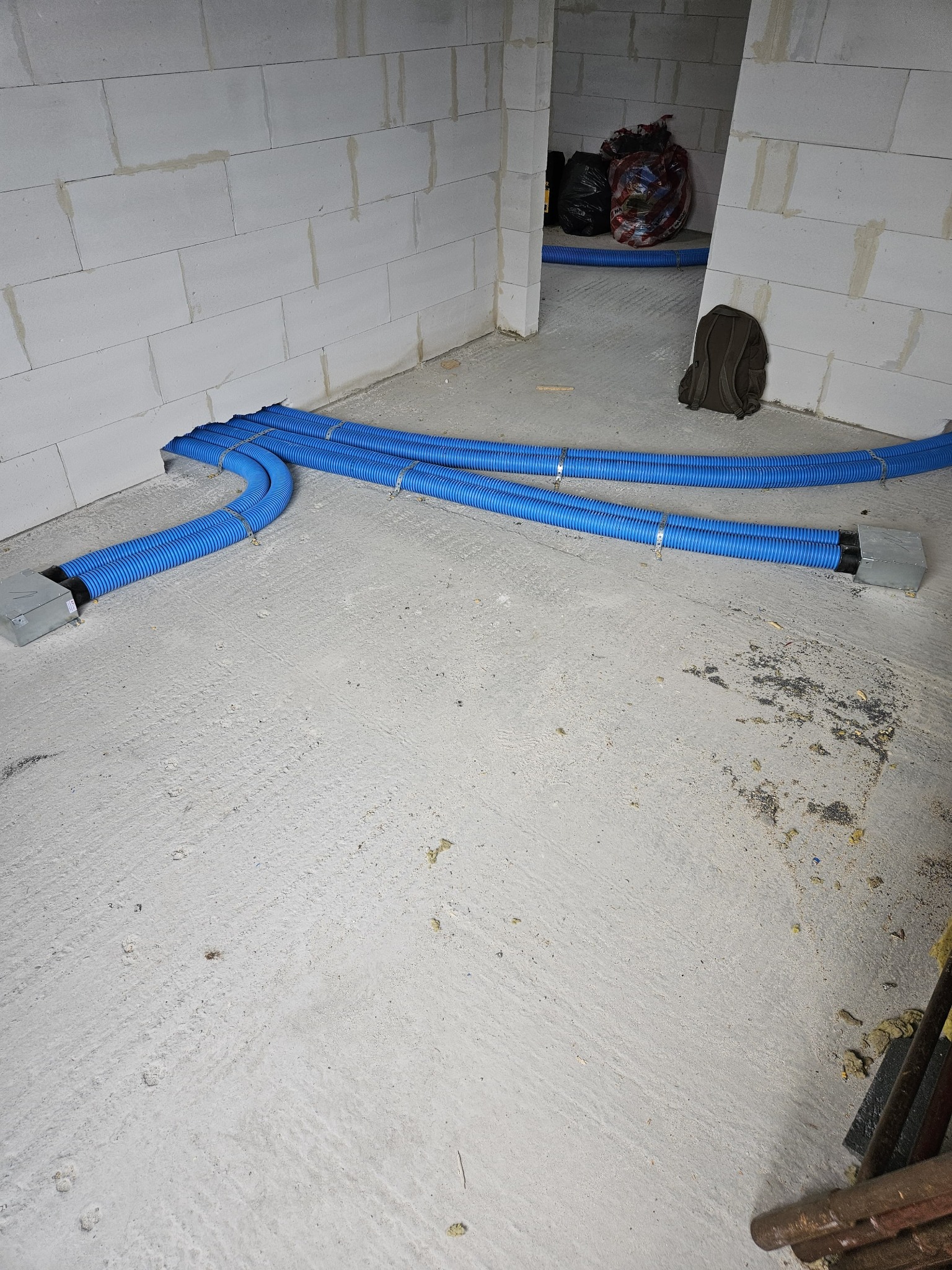 Niebieskie, karbowane rury wentylacyjne prowadzone po surowym betonowym podłożu wewnątrz budynku w stanie deweloperskim, z widocznymi elementami mocującymi i szarą puszką przyłączeniową.