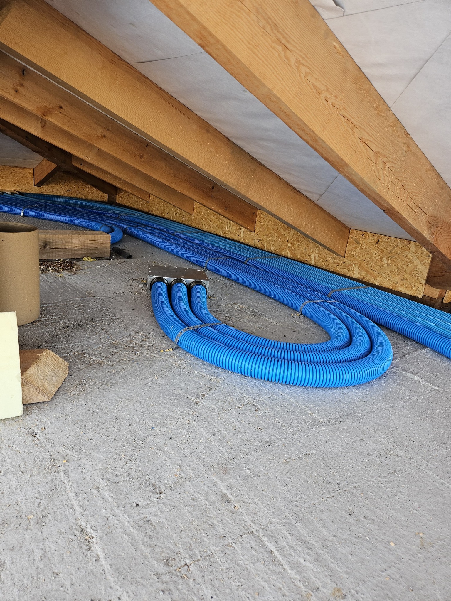 Niebieskie, karbowane rury wentylacyjne ułożone pod drewnianą konstrukcją dachu na surowym podłożu, widoczny fragment skrzynki rozdzielczej.