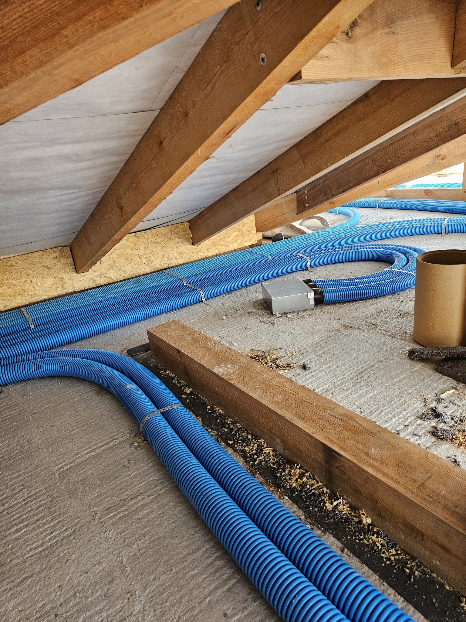 Niebieskie, karbowane rury wentylacyjne ułożone na betonowej podłodze poddasza, widoczne drewniane elementy konstrukcji dachu i izolacja.