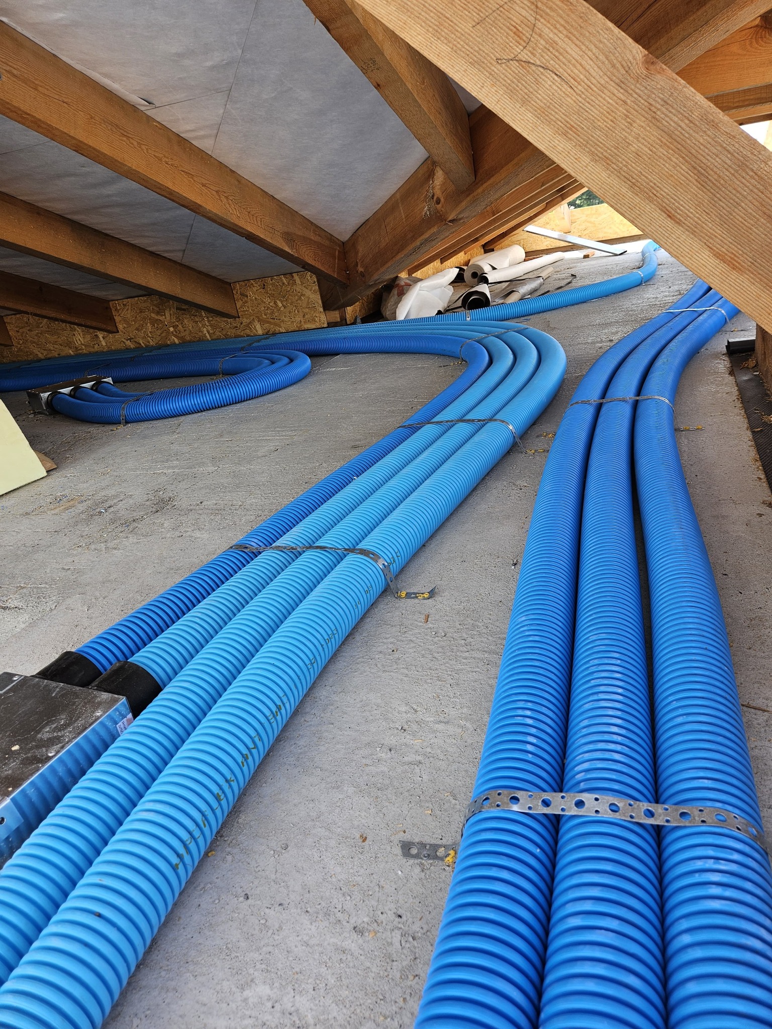 Niebieskie, karbowane rury wentylacyjne ułożone równolegle na betonowej podłodze poddasza, przymocowane metalowymi obejmami, widoczne drewniane krokwie dachu.