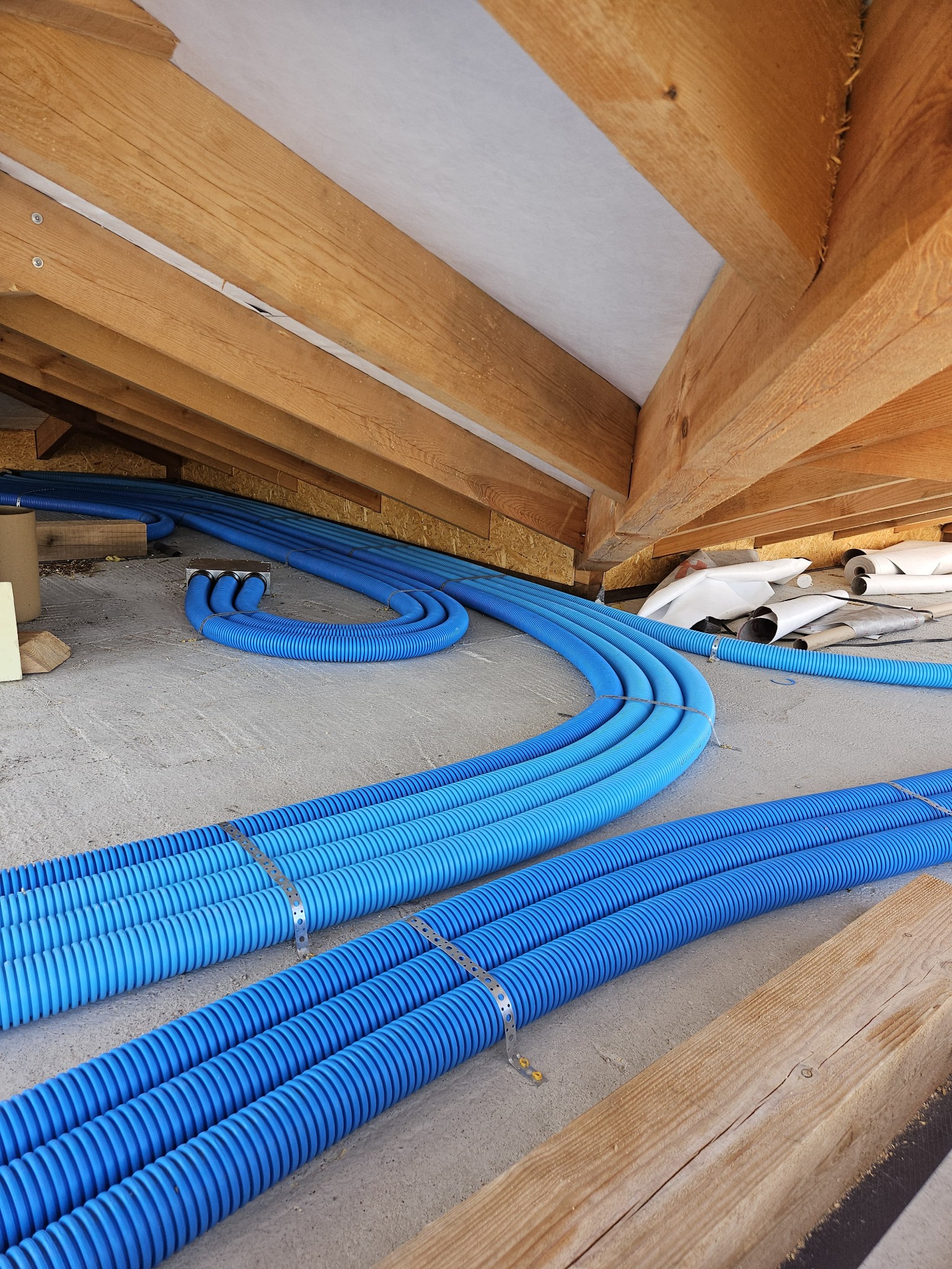 Niebieskie, karbowane rury wentylacyjne prowadzone pod drewnianą konstrukcją dachu, zabezpieczone metalowymi obejmami, z widocznymi materiałami izolacyjnymi w tle.