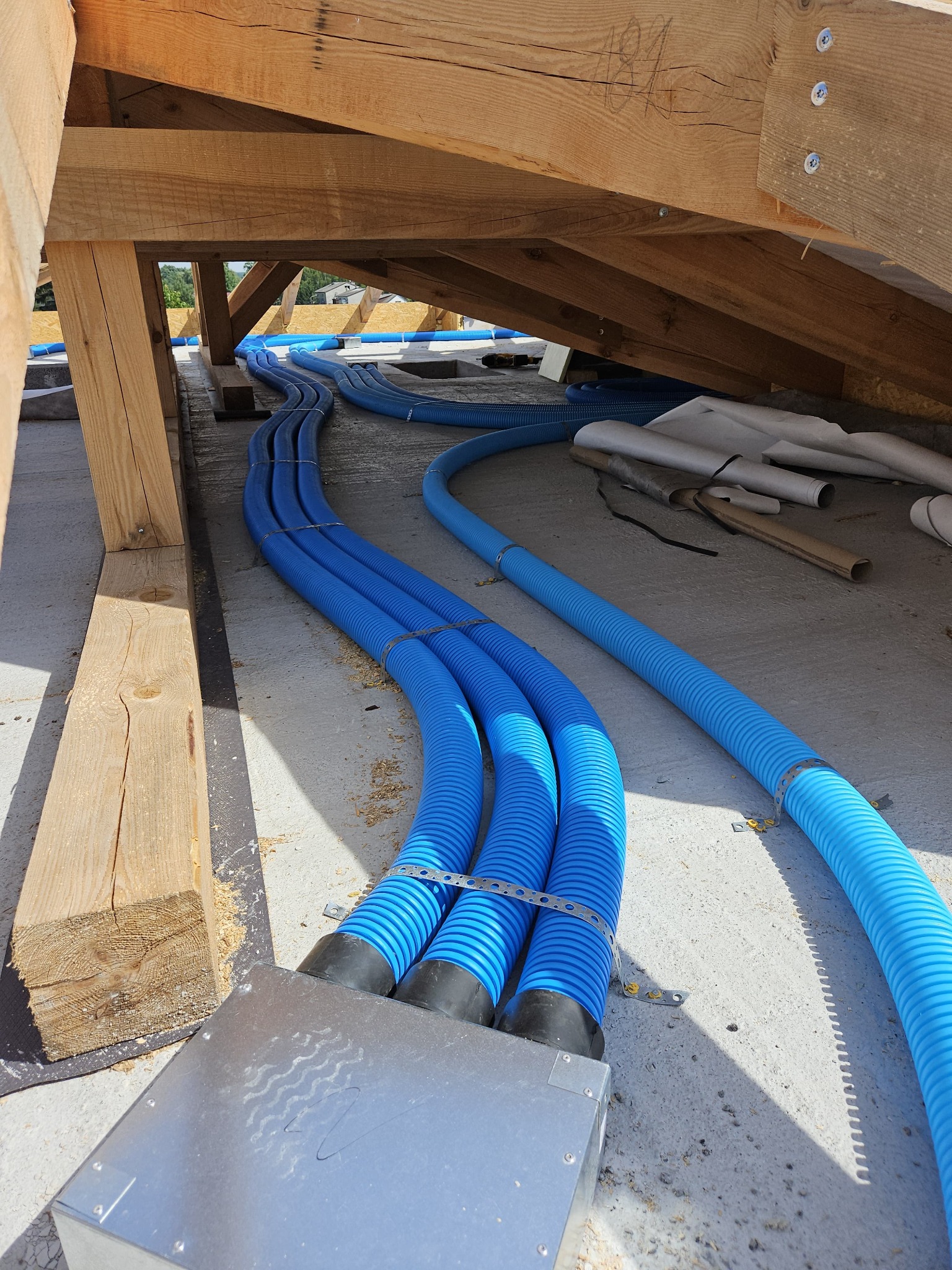 Niebieskie, karbowane przewody wentylacyjne przymocowane do metalowej skrzynki i drewnianej konstrukcji dachu na poddaszu, widoczne elementy montażowe i resztki materiałów budowlanych.