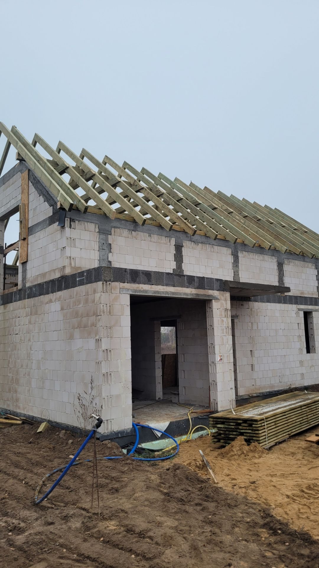 Surowa konstrukcja domu z białych bloczków, na której widać drewniany szkielet dachu w trakcie budowy, z niebem w tle.