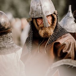 Photo & Brand - Fotografia, Strony WWW - Portret brodatego mężczyzny w hełmie wikinga z kolczugą, na tle innych wojowników. Ujęcie z bliska, skupione na twarzy i detalu stroju.