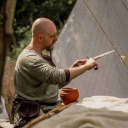 Photo & Brand - Fotografia, Strony WWW - Mężczyzna w stroju historycznym rzeźbi w drewnie nożem. Widoczny gliniany garnek i skórzana torba. W tle namiot i zieleń.